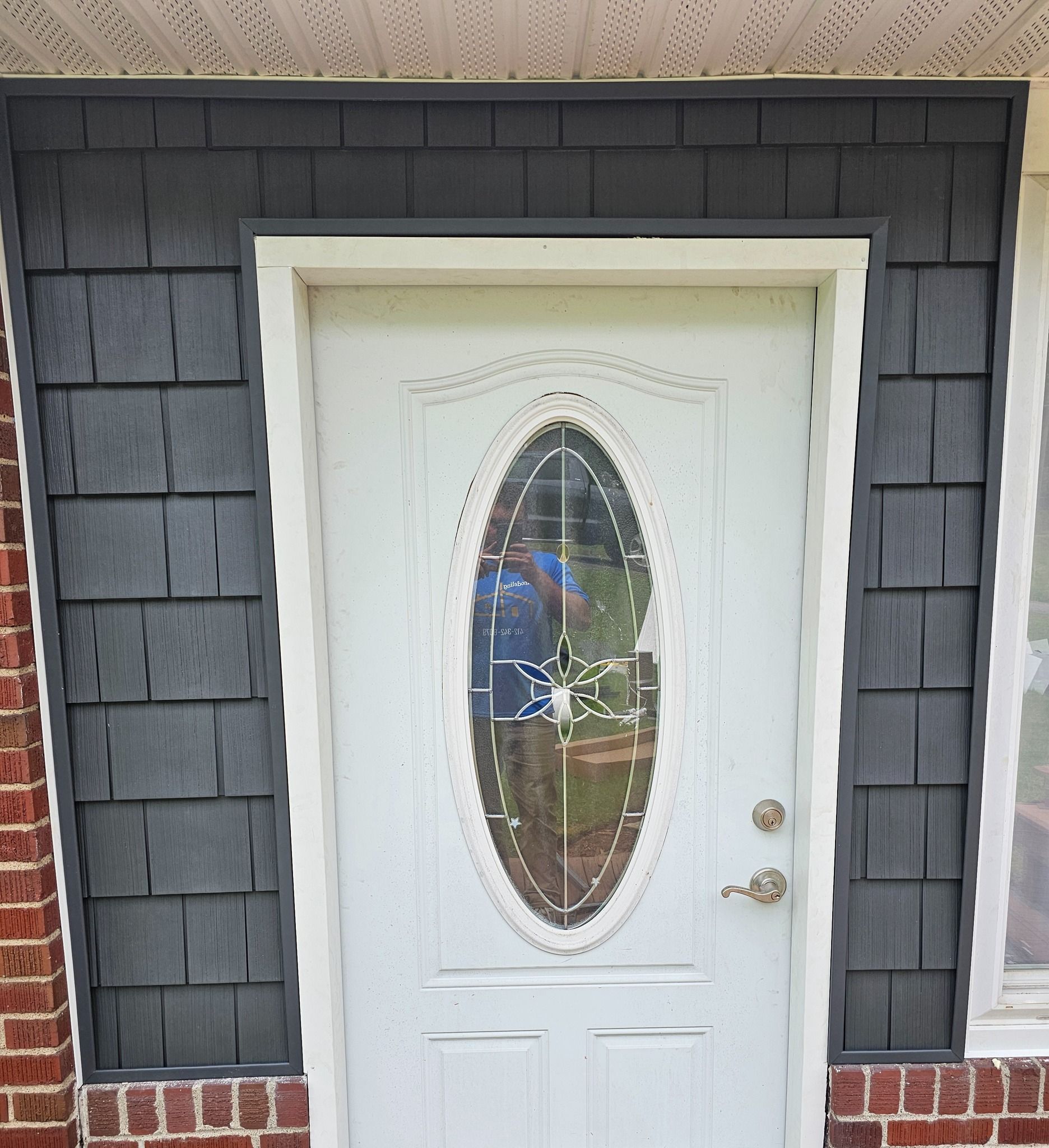 White door with oval window, framed by gray shingle siding.  Reflective surface shows a person.
