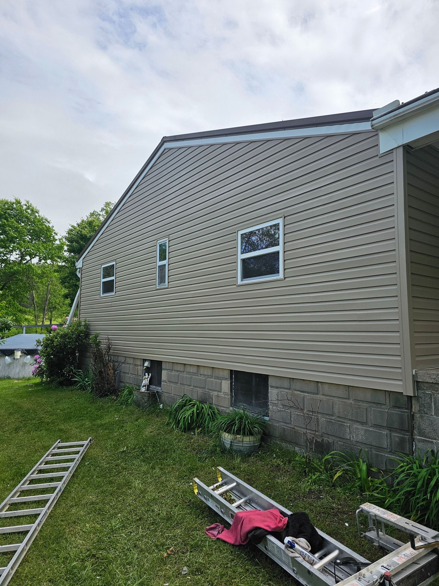 Side view of a house with light tan siding, a brown roof, and a gray stone foundation. 