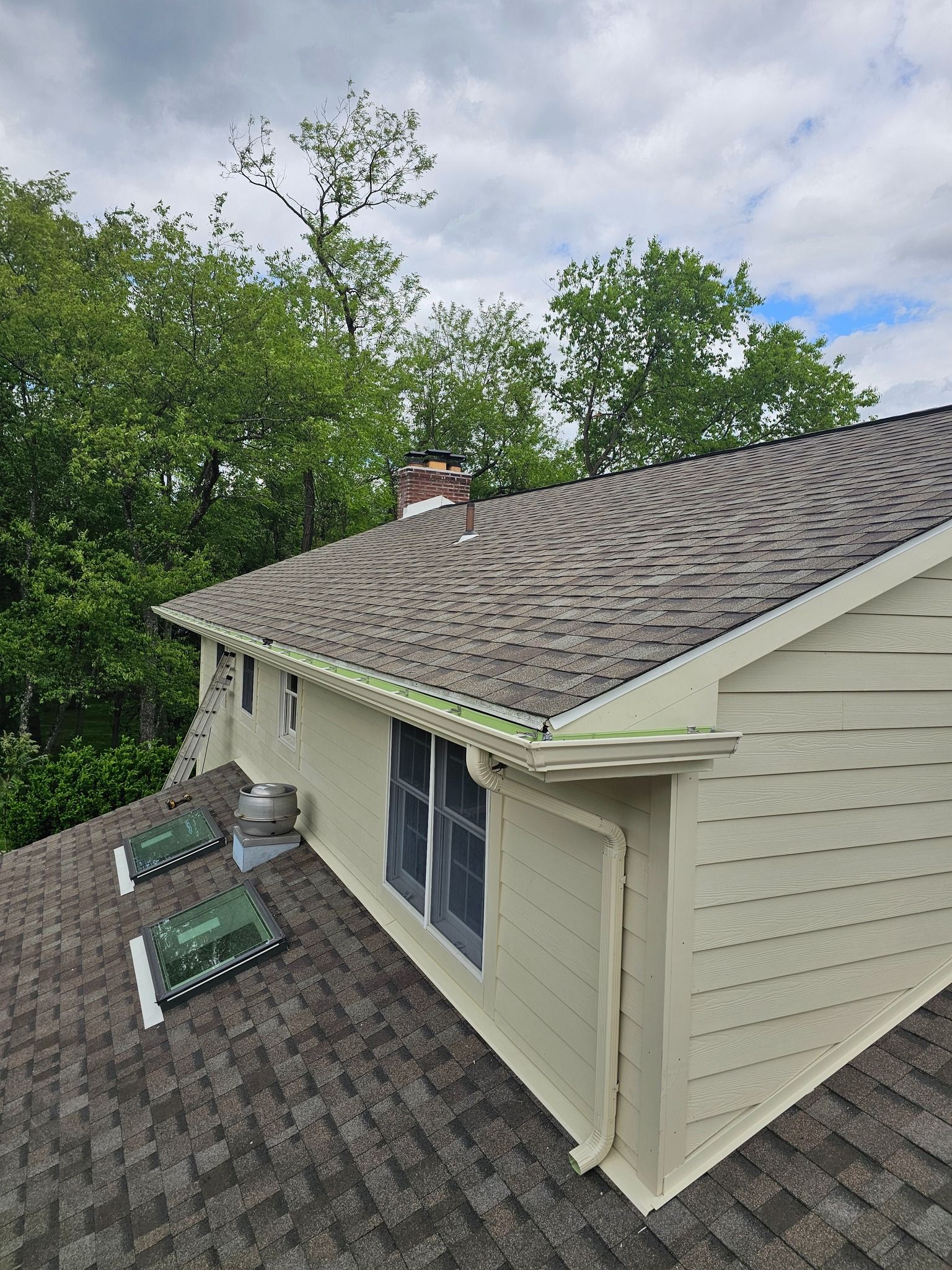 Overhead view of a house with a brown shingled roof, light siding, and green gutters under a cloudy sky.
