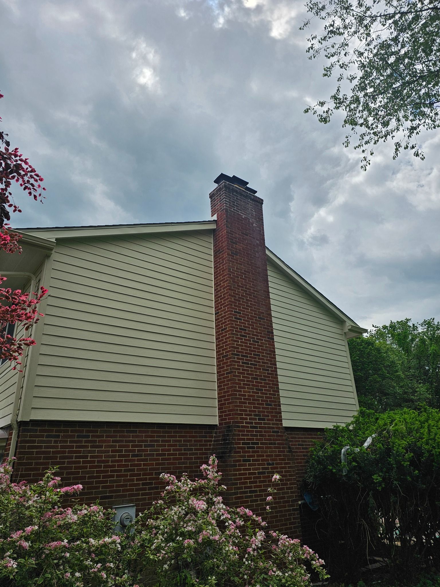 Brick chimney on house with white siding under a cloudy sky, surrounded by greenery.