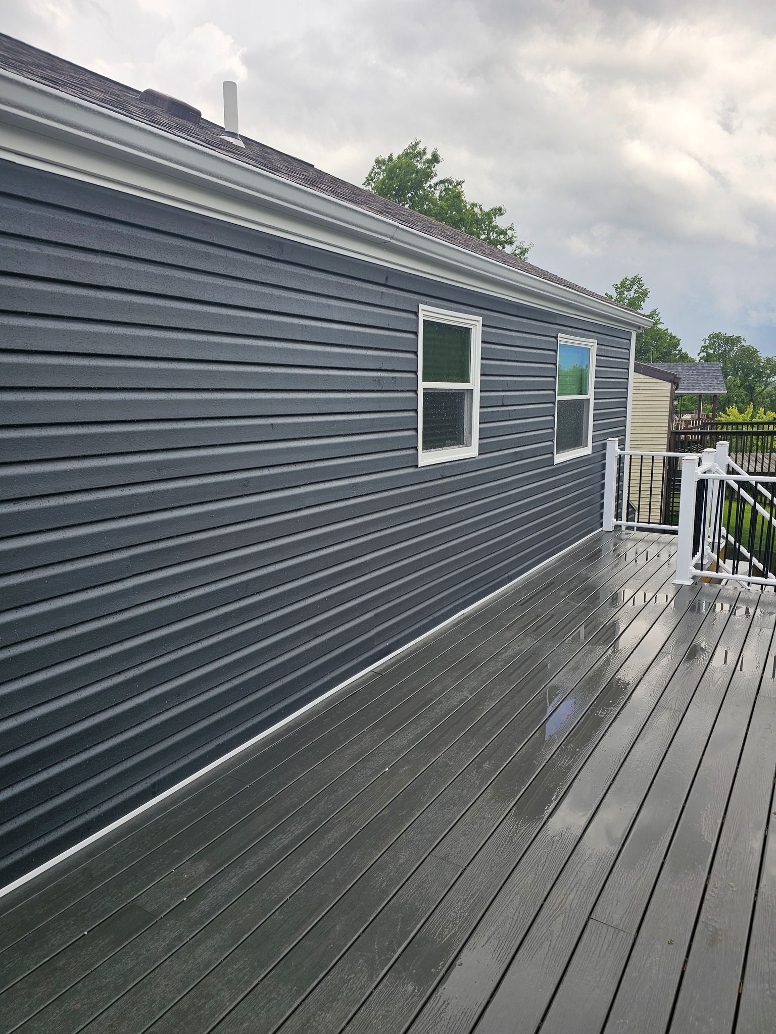 Dark blue siding on a house next to a gray wooden deck, under a cloudy sky.