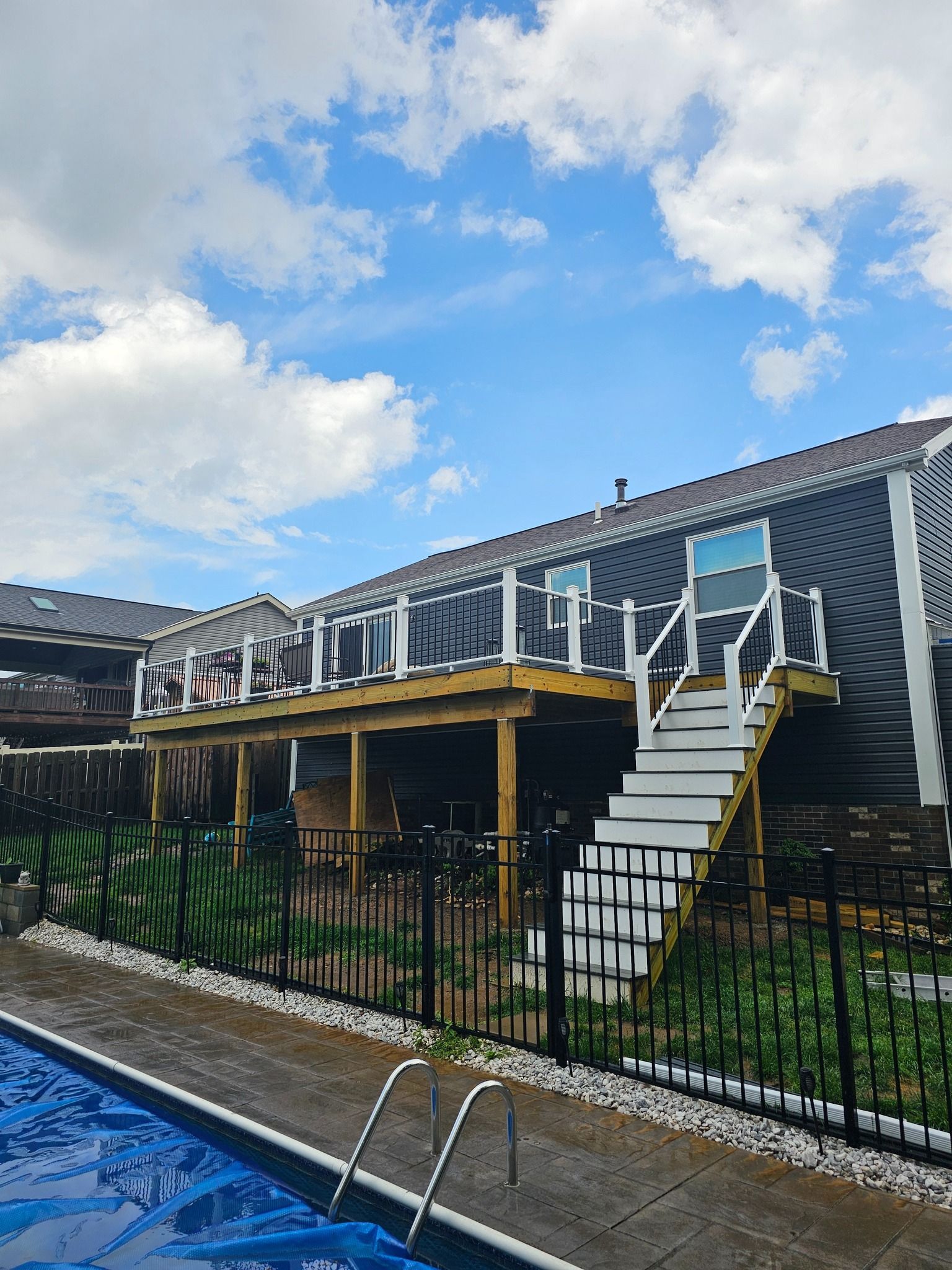 Backyard with pool and elevated wooden deck with stairs. Black fence surrounds pool and deck. Blue sky.
