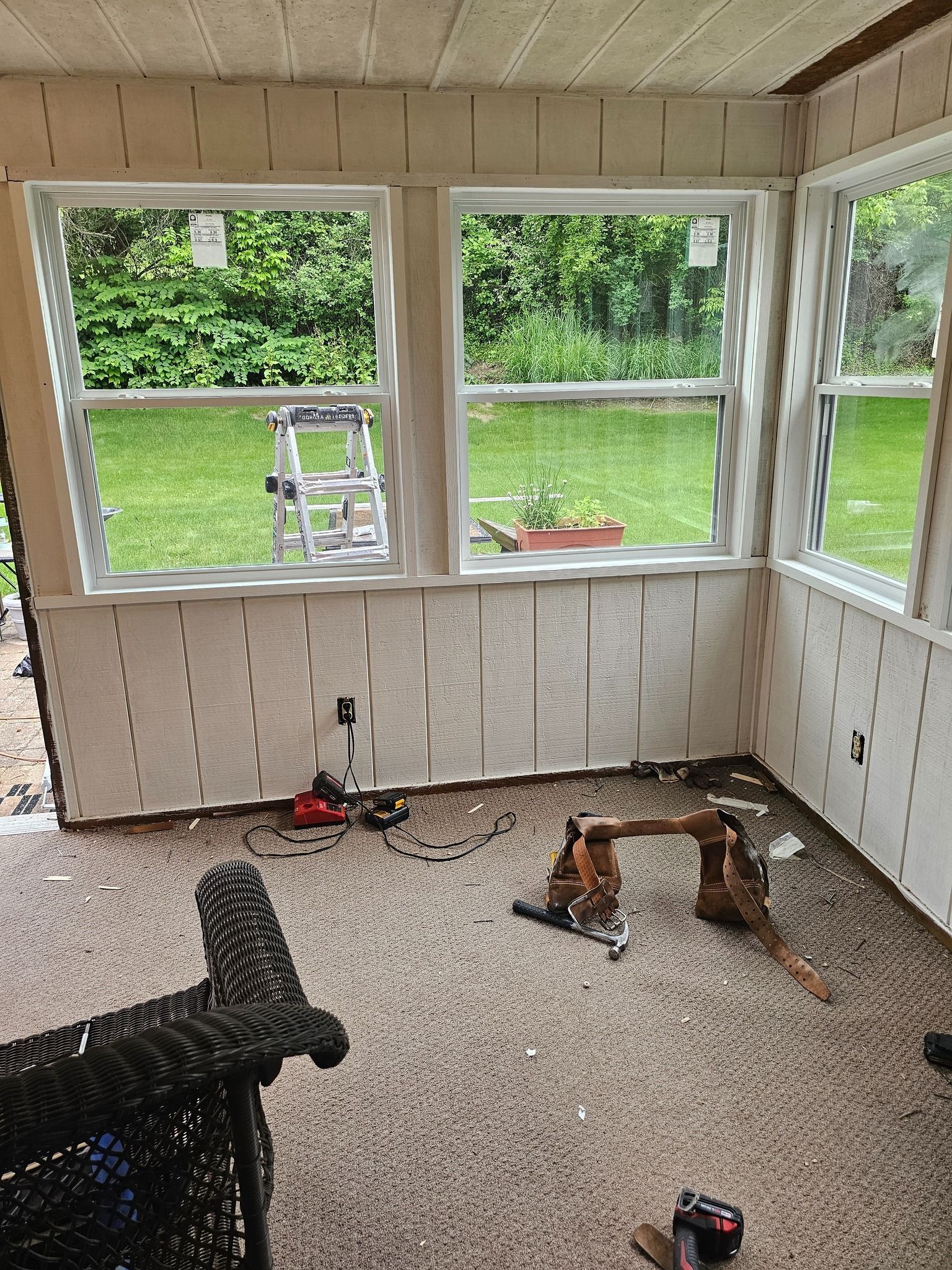 Sunroom with new windows, light-colored paneling, and a woven chair. Tools and carpet are visible.