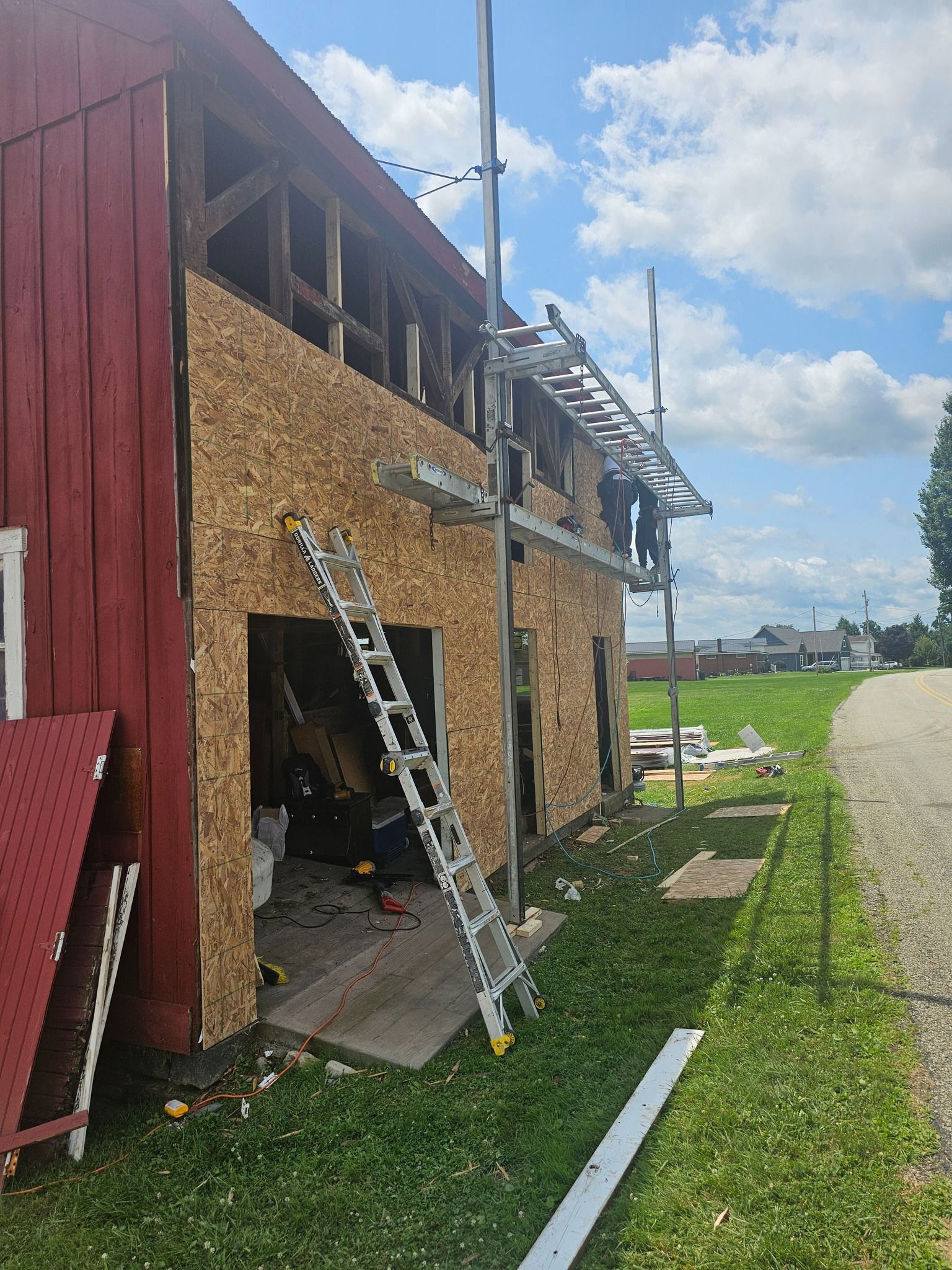 Barn construction: Workers on scaffolding, installing siding. Red barn, plywood siding, ladder, green grass, blue sky.