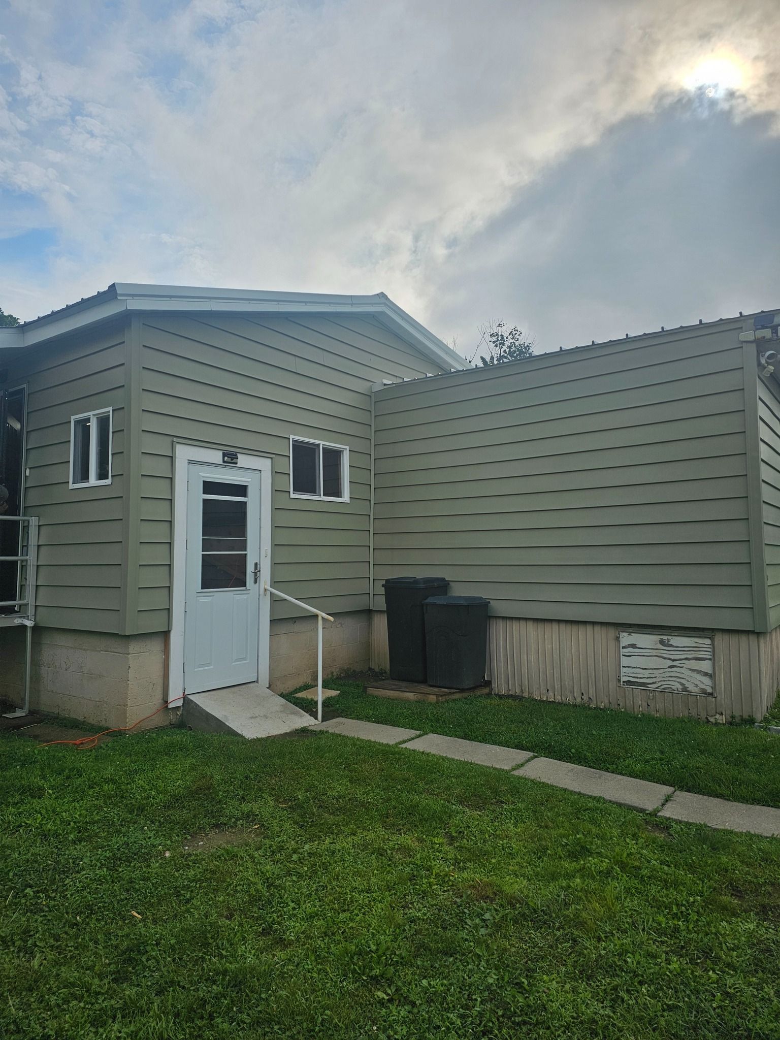 Green-sided building with white door and windows, concrete ramp, two black bins, and a grassy yard.