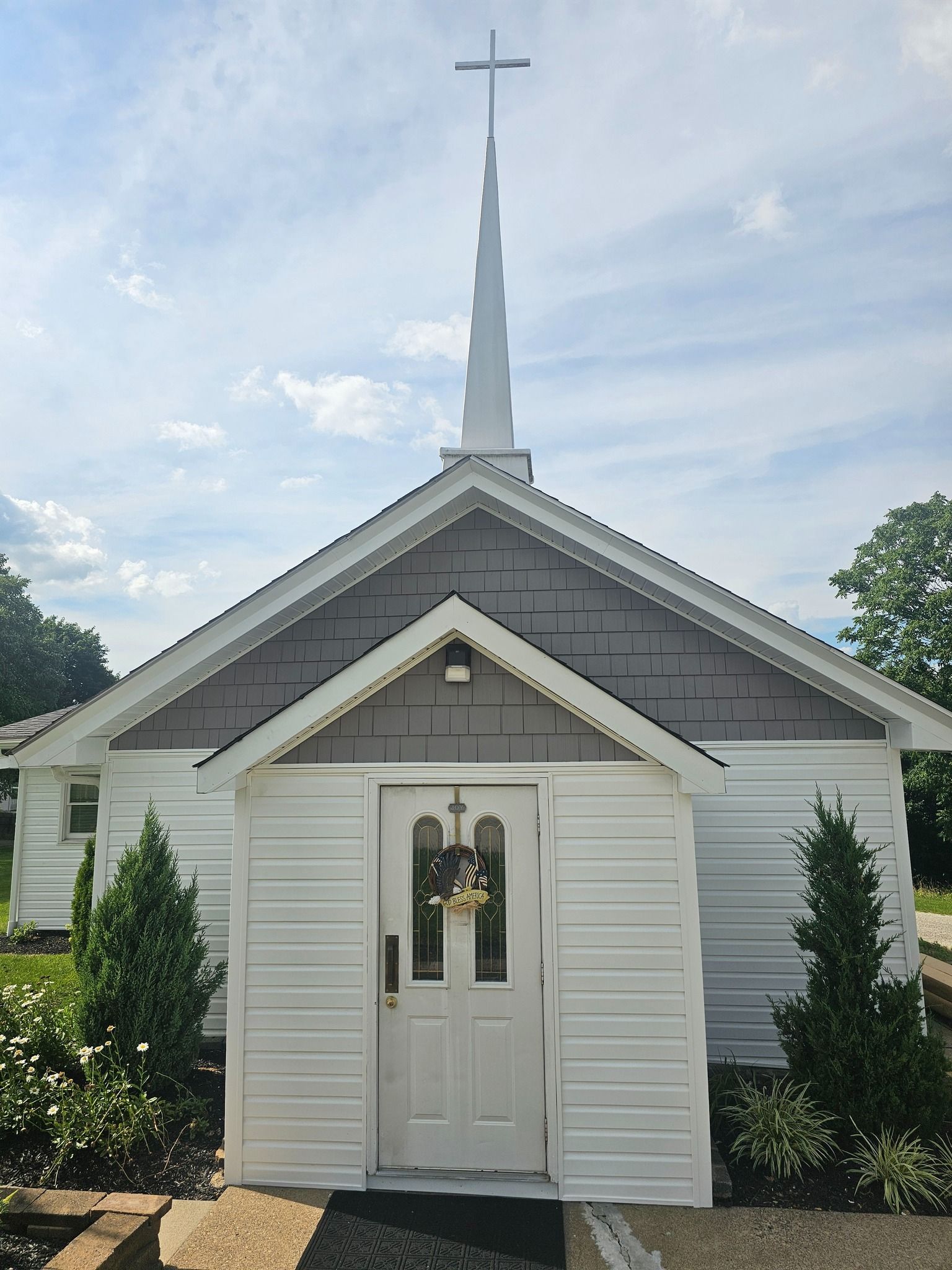 White church with a steeple, cross, and entry porch. Green shrubs flank the door.