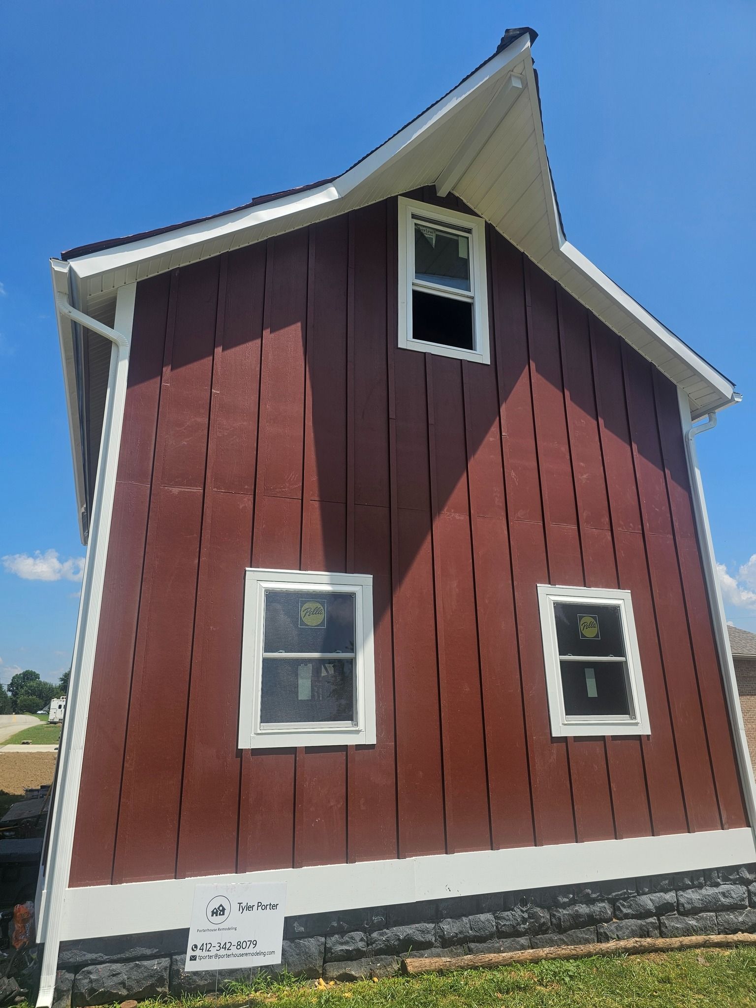 Red barn-style building with white trim, three windows, and a dark stone foundation against a blue sky.