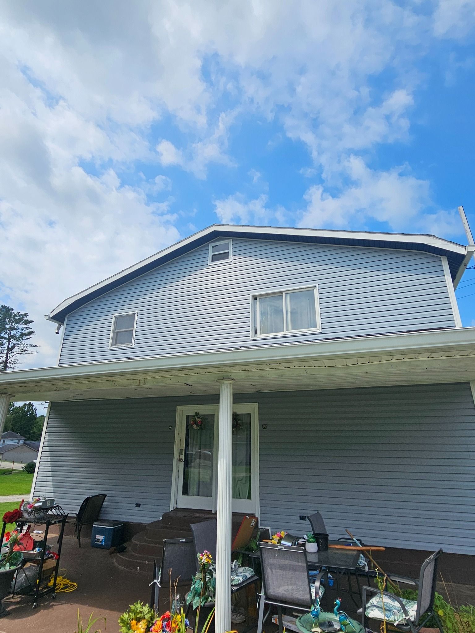 Two-story house with gray siding, a white awning, and a blue sky. Chairs and tables are under the awning.