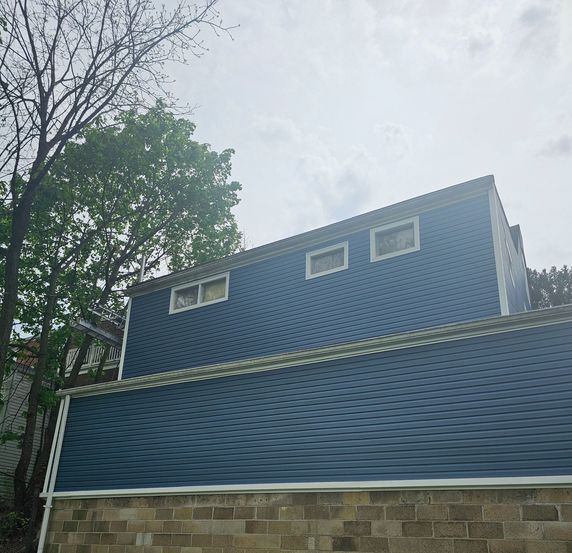 Blue-sided house with three rectangular windows on upper level. Green tree in the background. Cloudy sky.