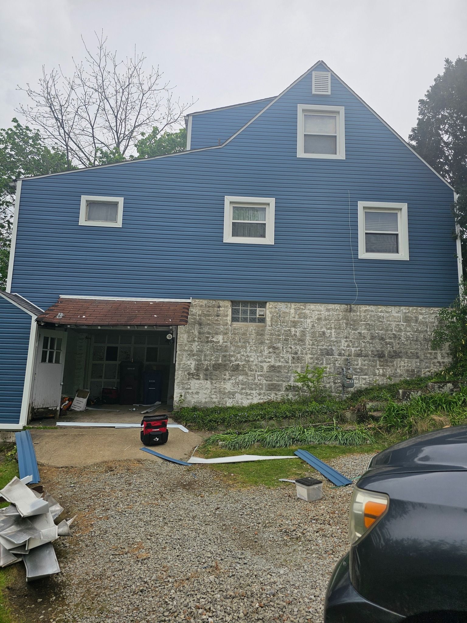 Blue-shingled house exterior; garage with exposed concrete; gravel driveway with car parked; overcast sky.