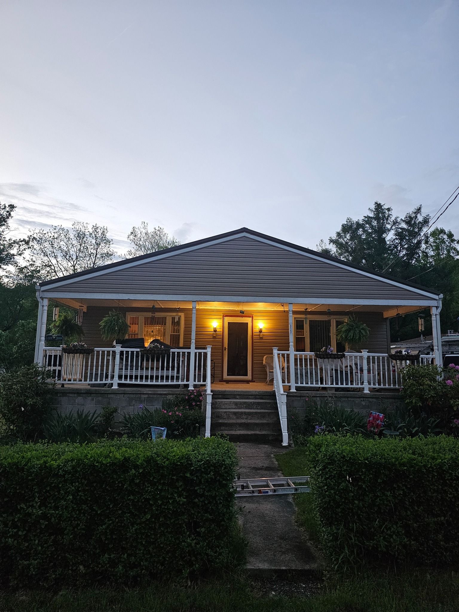 Cottage with porch lit by warm lights. White railings and steps lead to front door. Bushes and trees surround.