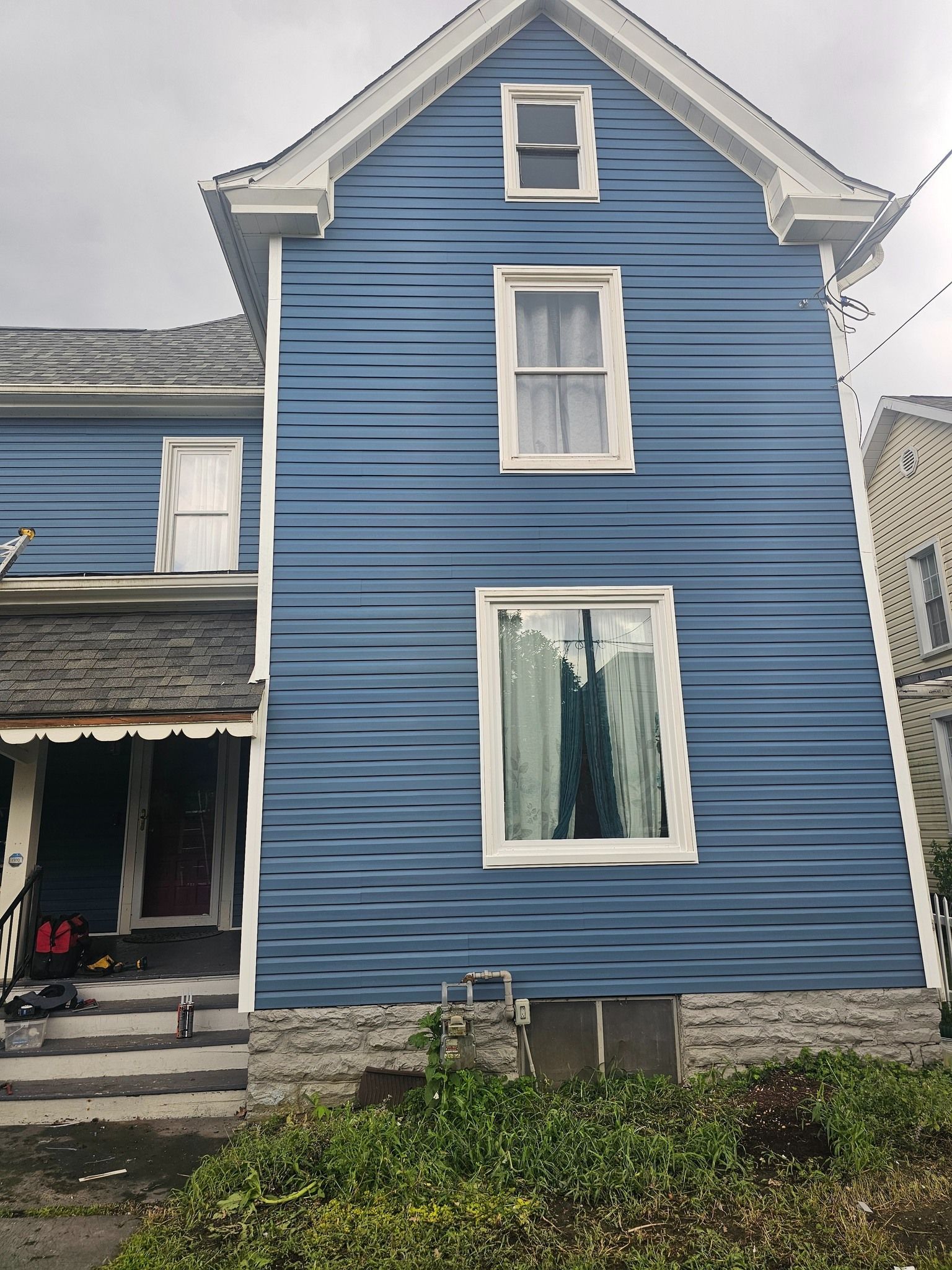 Blue two-story house with white trim and windows. Green bushes in front. Gray sky overhead.