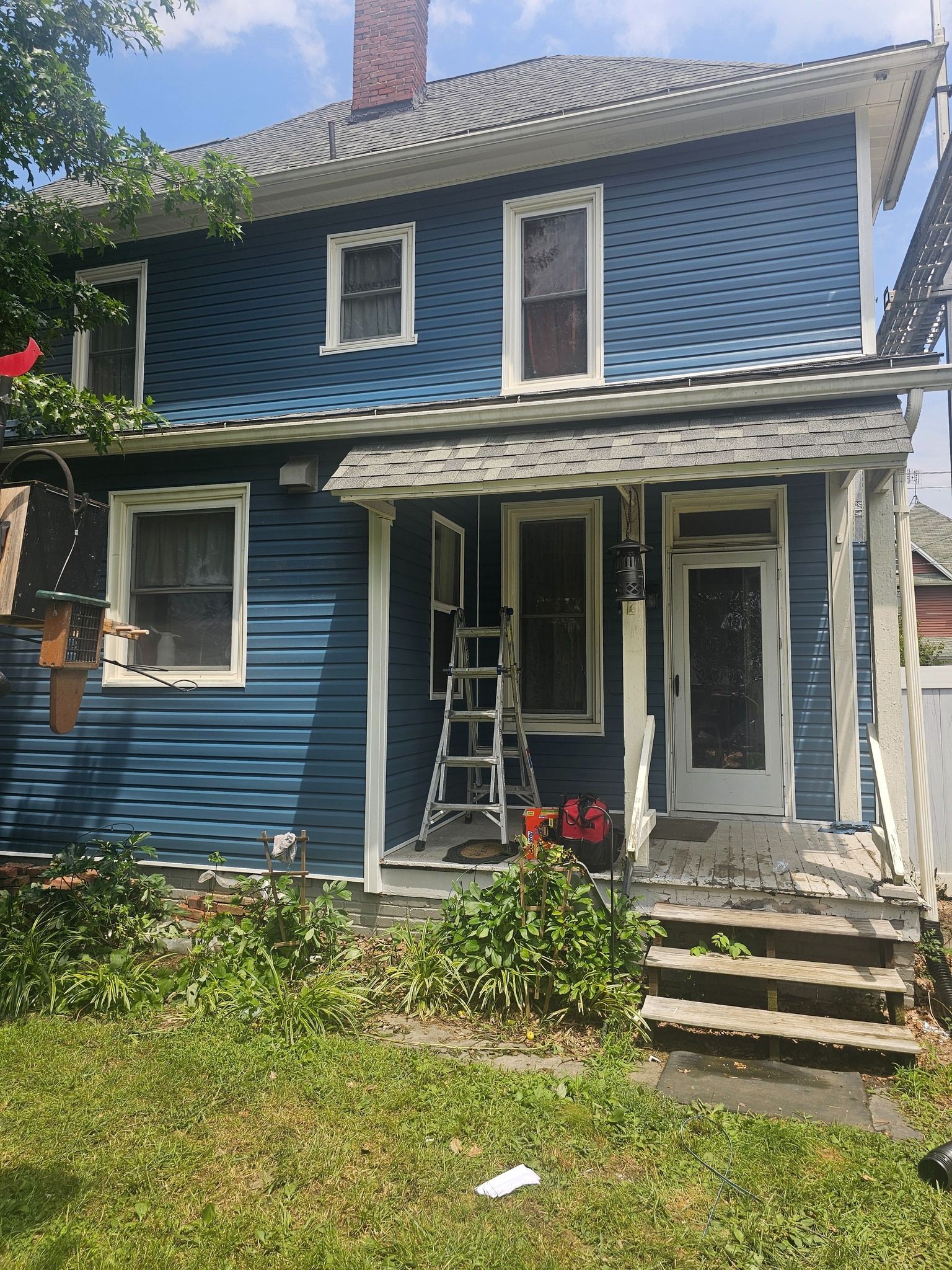 Blue two-story house with overgrown yard and porch. A ladder is leaning against the door.