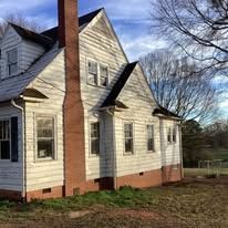 Weathered white house with red brick chimney and overgrown yard.