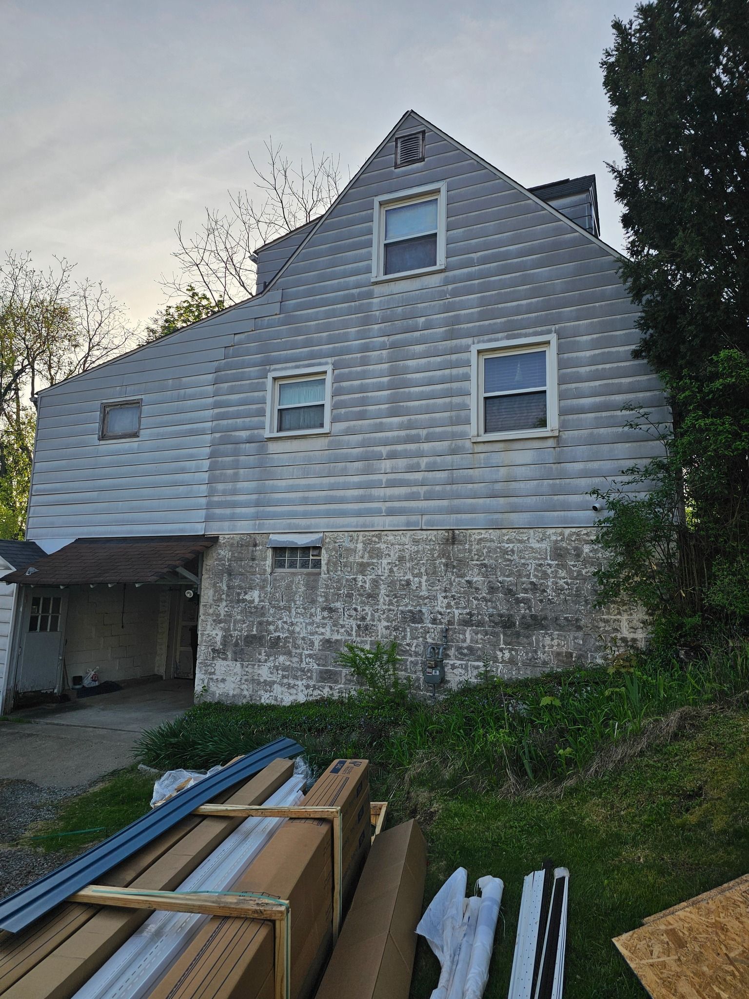 Back of a two-story house with gray shingle siding, a carport, and construction materials on the ground.
