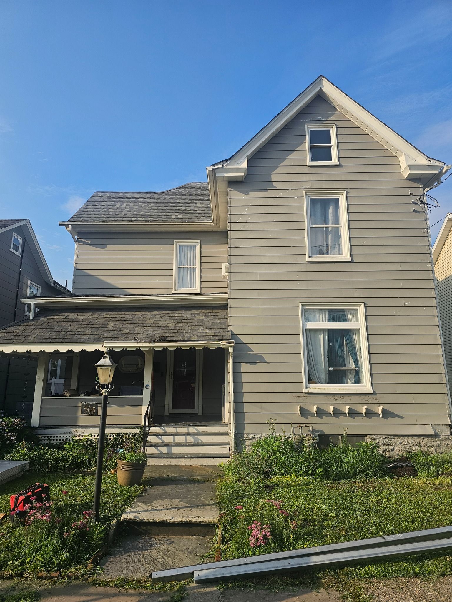 Two-story gray house with porch and overgrown front yard, under a blue sky.