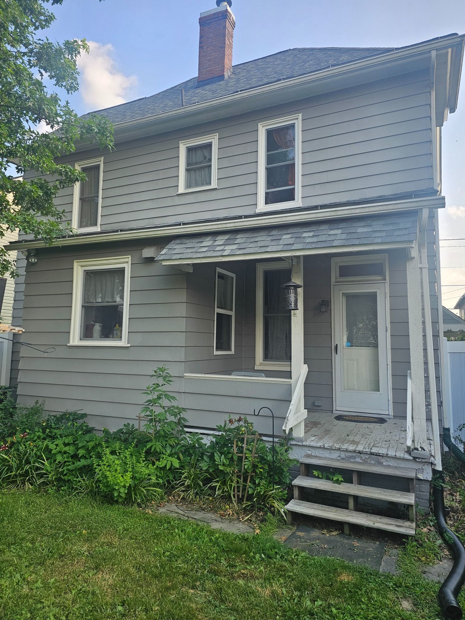 Two-story gray house with white trim. Porch with steps. Overgrown bushes in front yard.