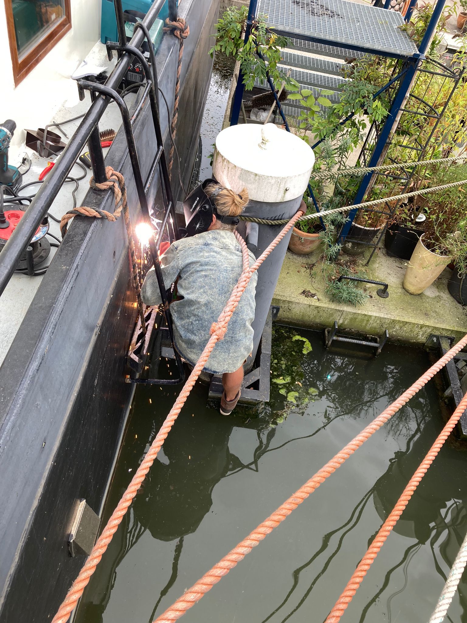 Een persoon last aan een zwarte metalen constructie naast een waterpartij; touwen en planten zijn zichtbaar.