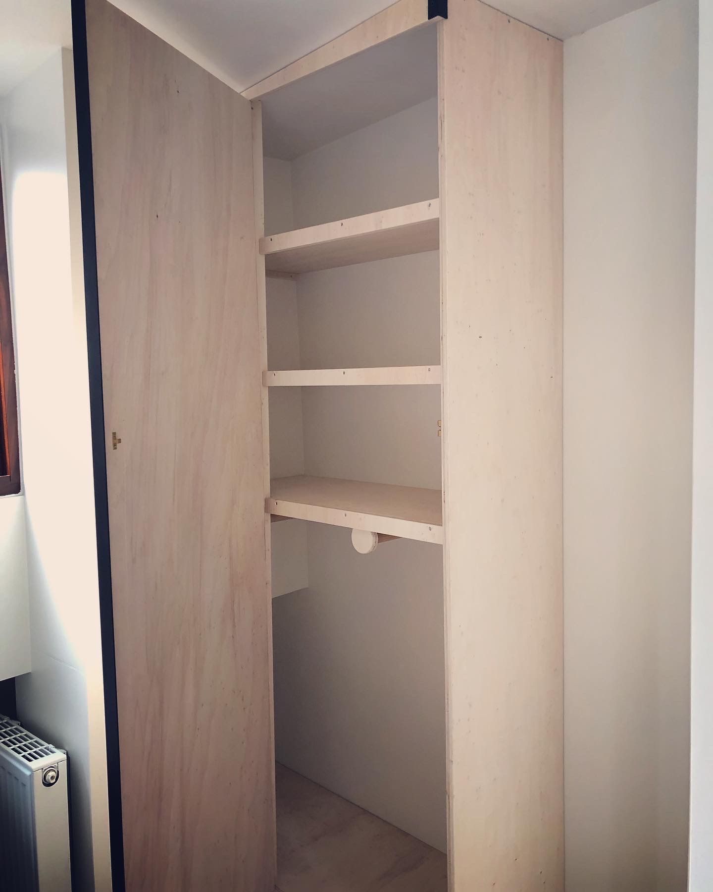 Een ingebouwde houten kast met drie planken en een schuifdeur; de kast is licht van kleur en heeft een smalle opening.