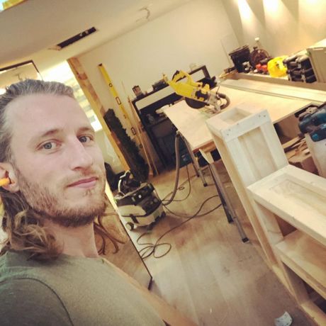 Man in werkplaats met oordoppen, omringd door houtbewerkingsgereedschap en houten werkstukken.