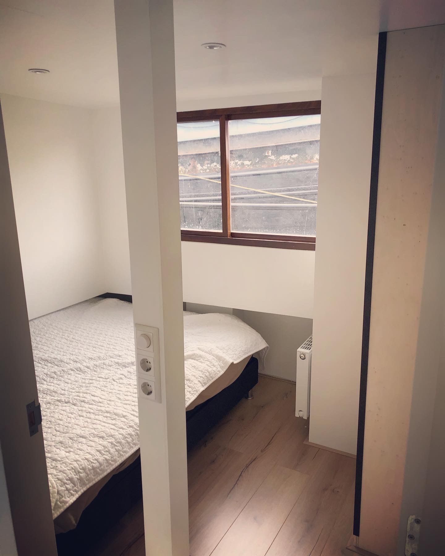 Kleine slaapkamer met een bed en een raam, zichtbaar vanuit een deuropening. Beige muren en houten vloer.