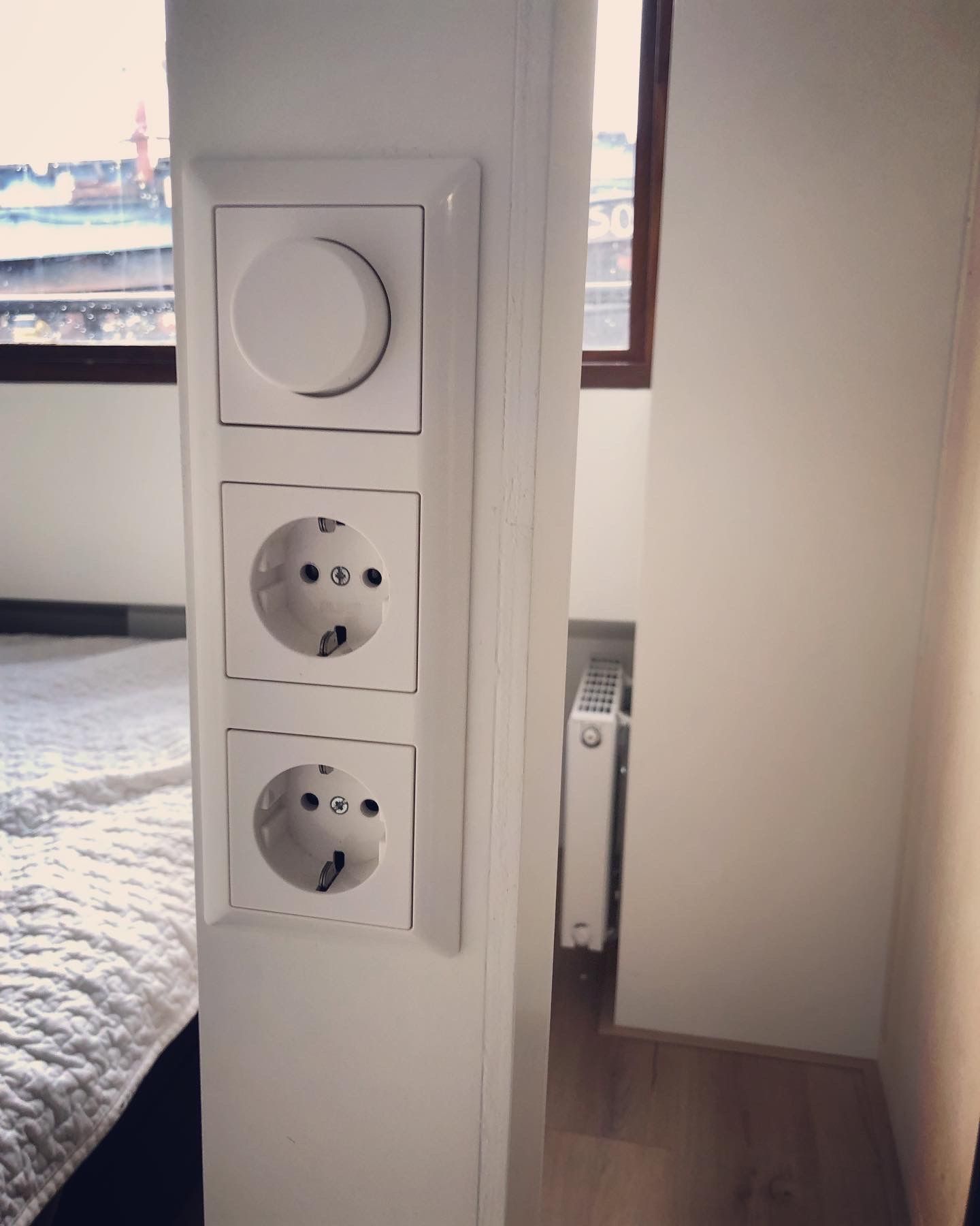 Witte stopcontacten en een dimmer op een witte muur, naast een raam en een kleine radiator.
