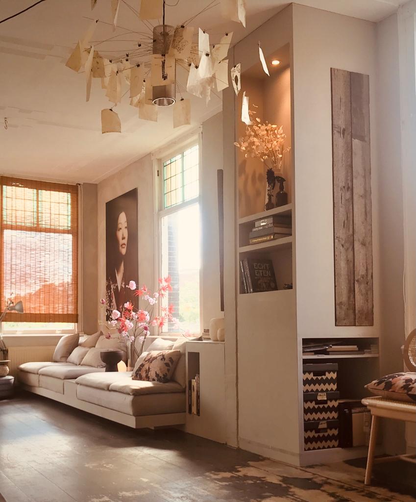 Woonkamer met crèmekleurige bank, kunstprint, ingebouwde planken en decoratieve lamp.