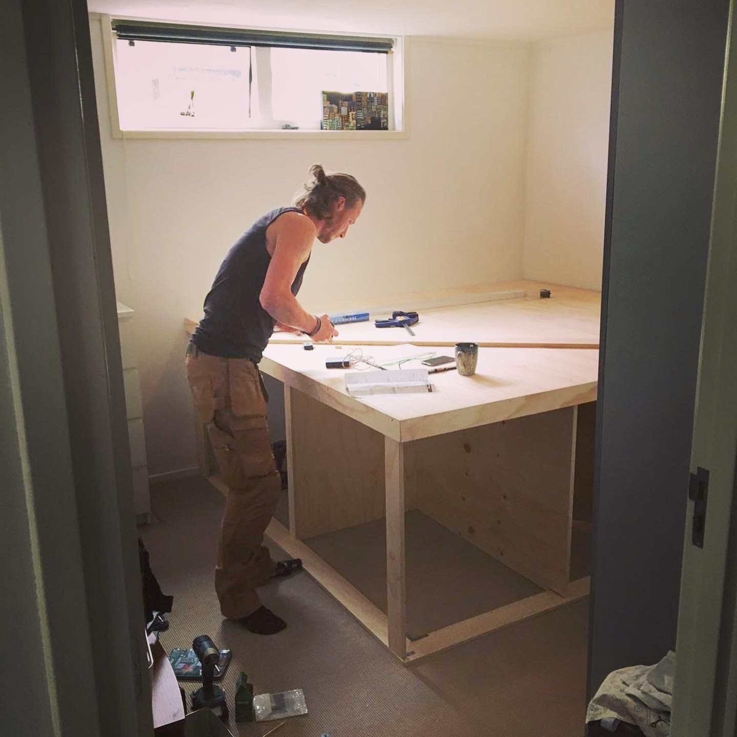 Een man, gekleed in een hemdje en cargobroek, is bezig een grote houten tafel in elkaar te zetten in een kamer met een raam.