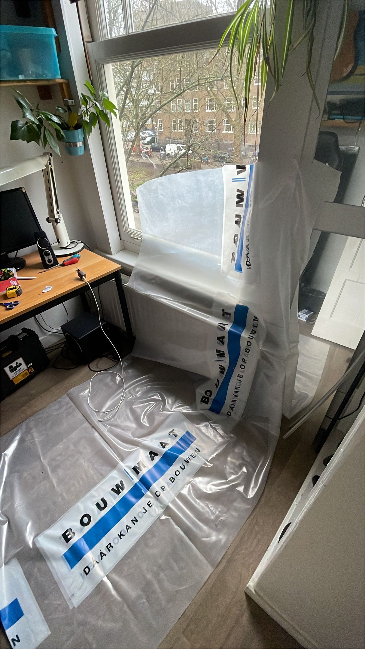 Plastic folie is met tape aan een raam bevestigd, bedekt de vloer en biedt een zonnig uitzicht naar buiten.