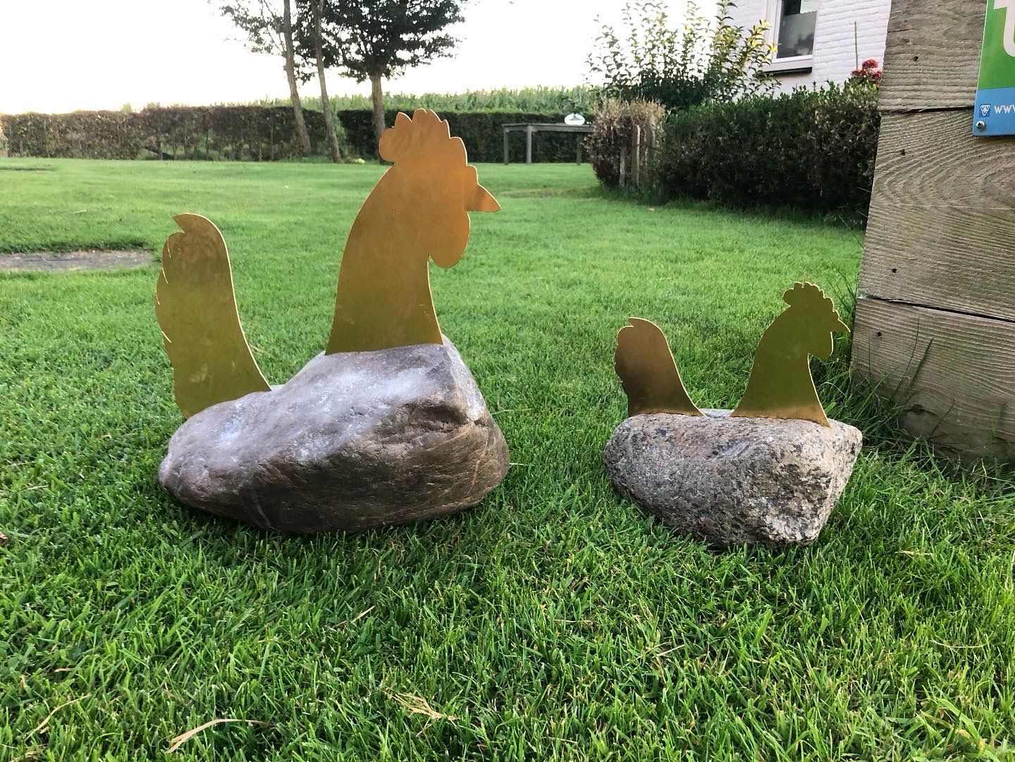 Twee metalen beelden van een haan en een hen op rotsen in een grasveld.