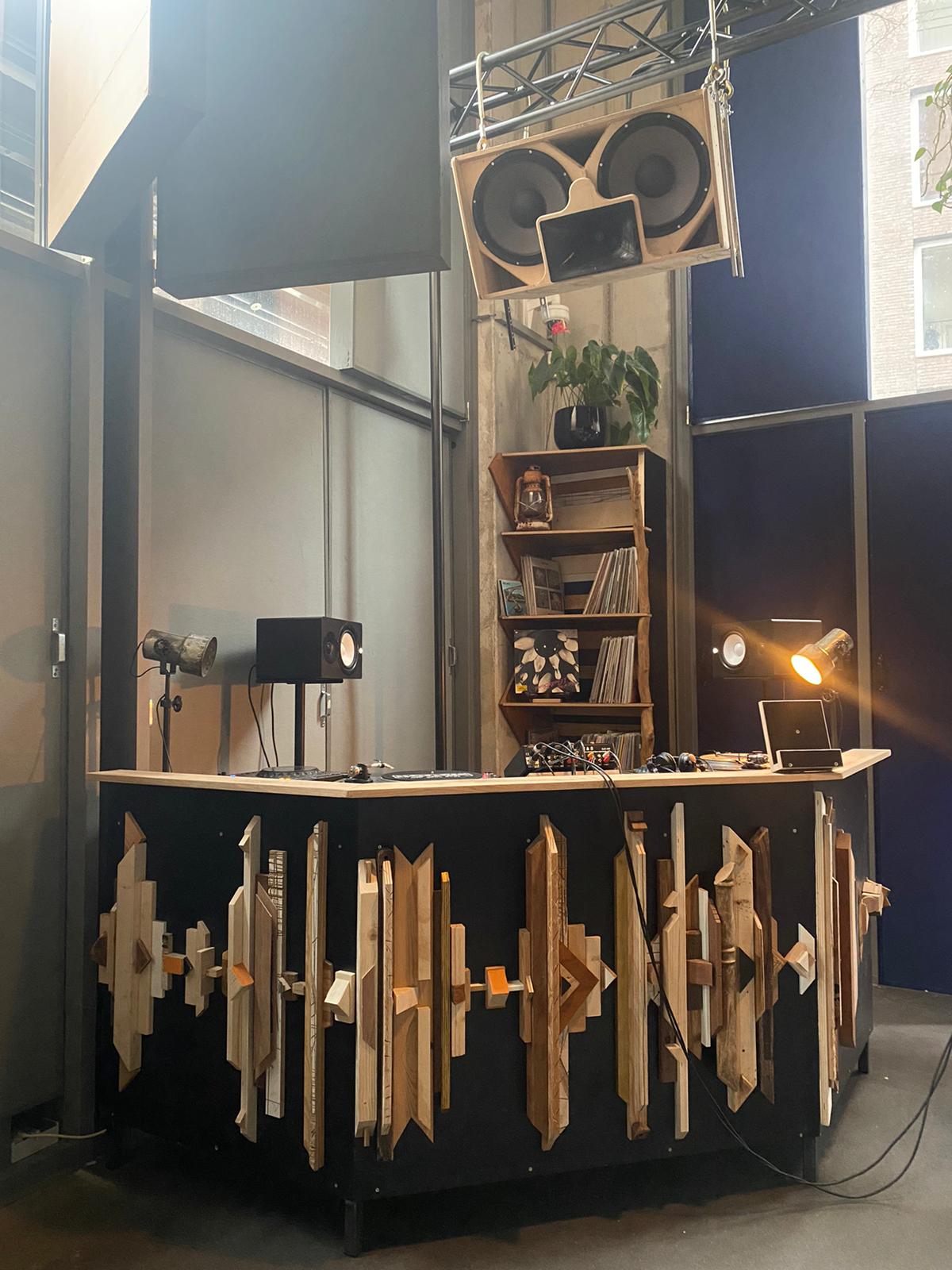 DJ-booth met geluidsgolfdesign, hangende speakers, planken met planten en platen, en een spotlight.