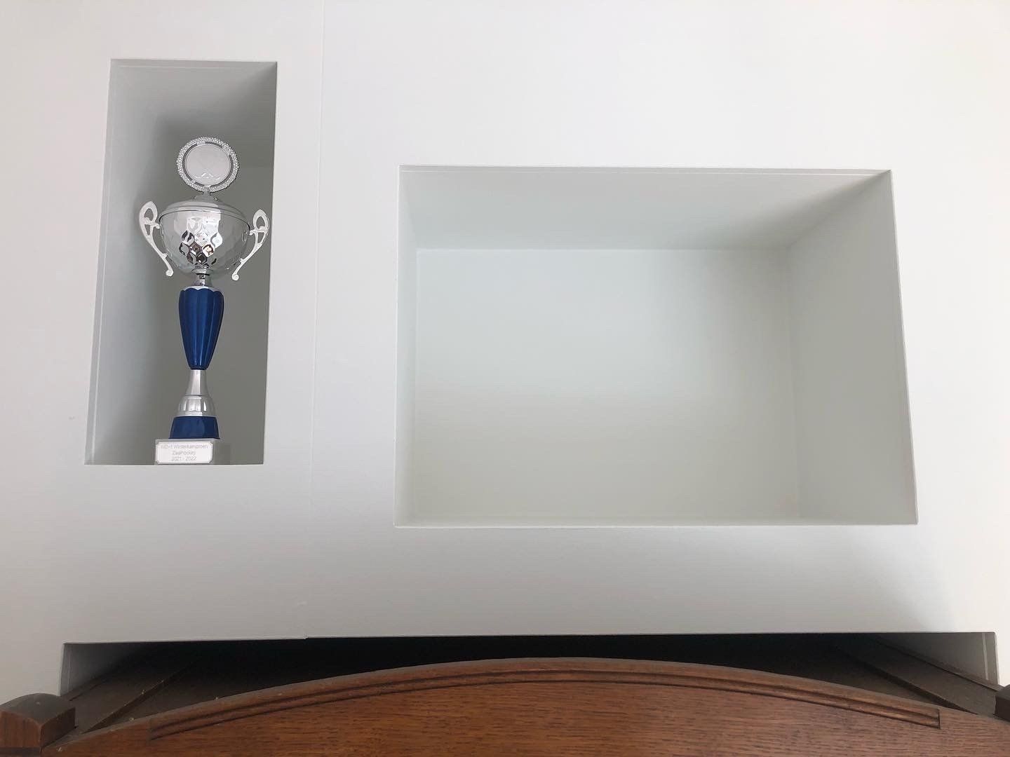 Witte muur met twee ingebouwde vierkante planken, waarvan er één een zilveren en blauwe trofee bevat.
