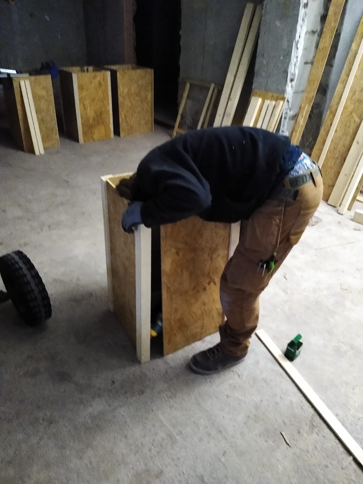 Iemand leunt tegen een houten kist, mogelijk om die in elkaar te zetten. Werkplaats met andere kisten, hout en een autoband.