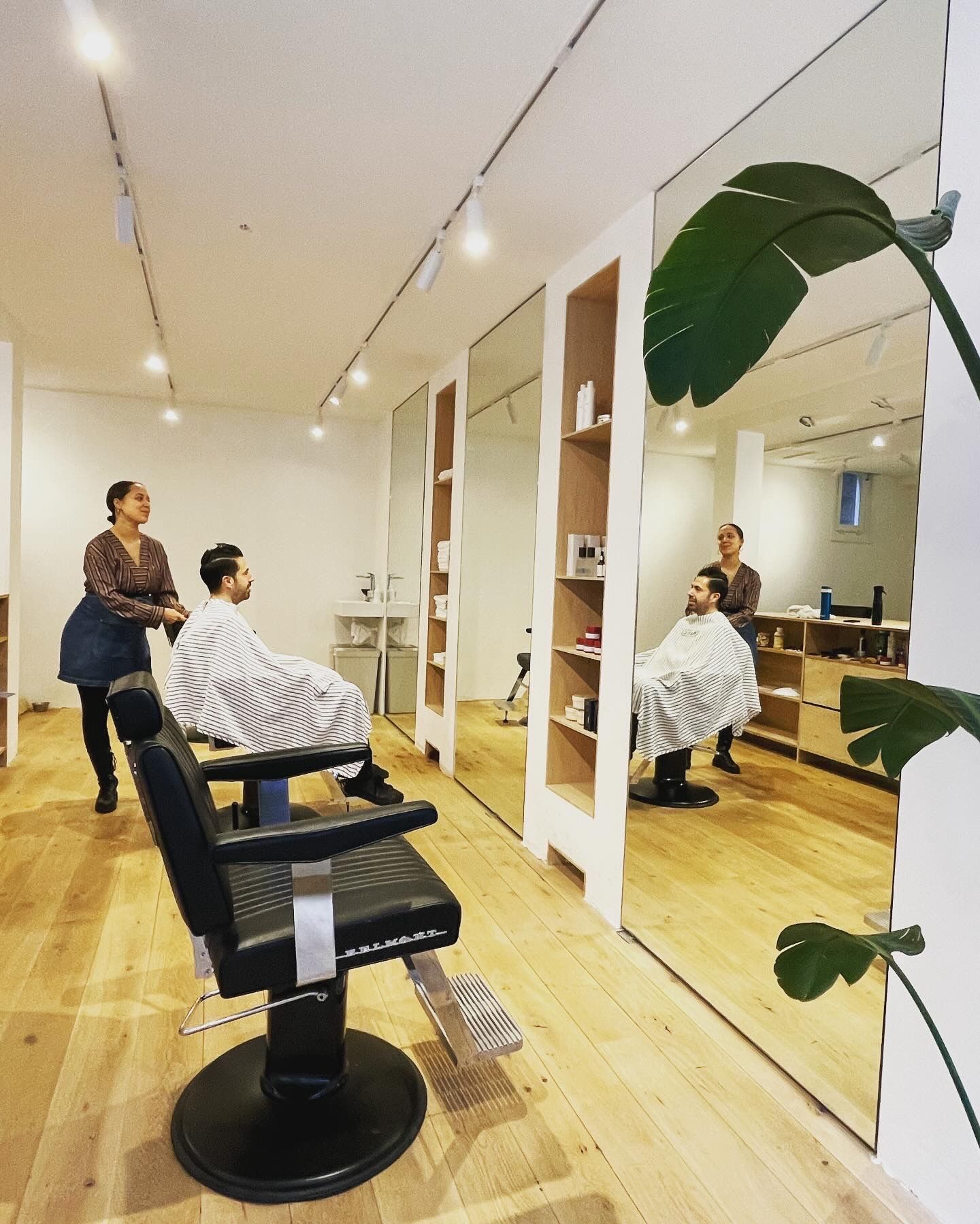 Interieur van een kapsalon: Twee kappers aan het werk bij klanten. Grote spiegels, houten vloer en een potplant.