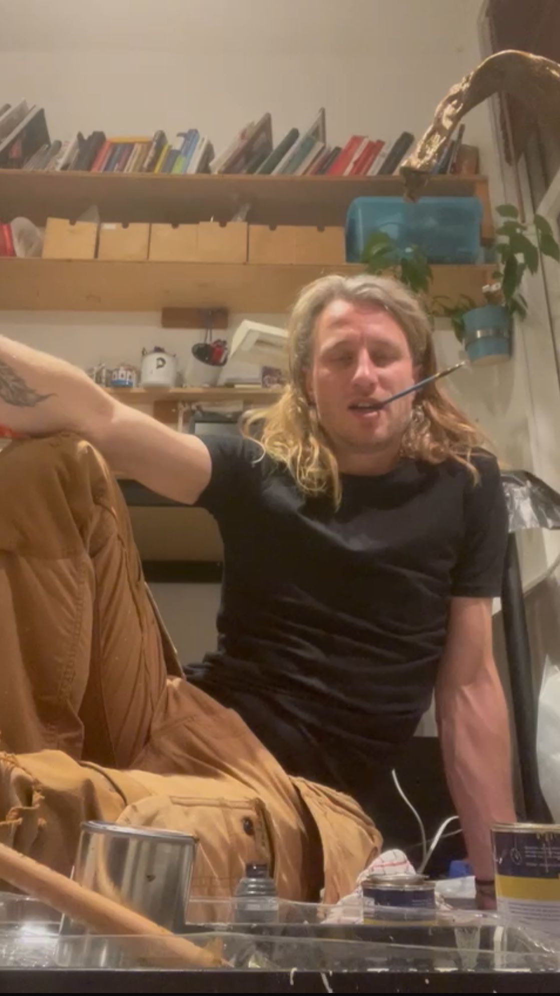 Een man met lang blond haar, een zwart shirt en een bruine broek zit er nonchalant bij en houdt een bril in zijn mond.