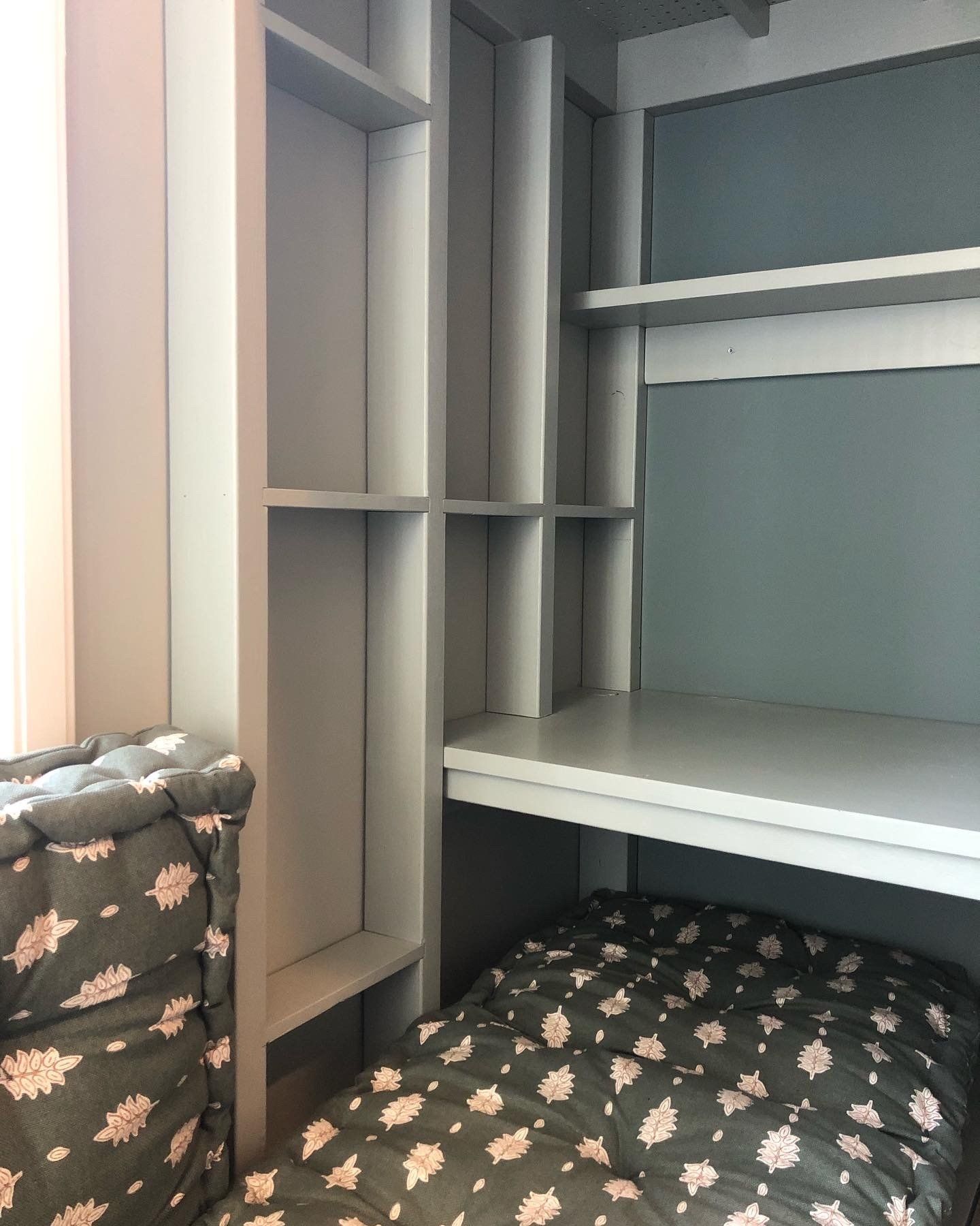 Ingebouwd bureau en planken in lichtgrijs met daaronder een bed. Grijs en zwart patroon beddengoed en stoelkussen.