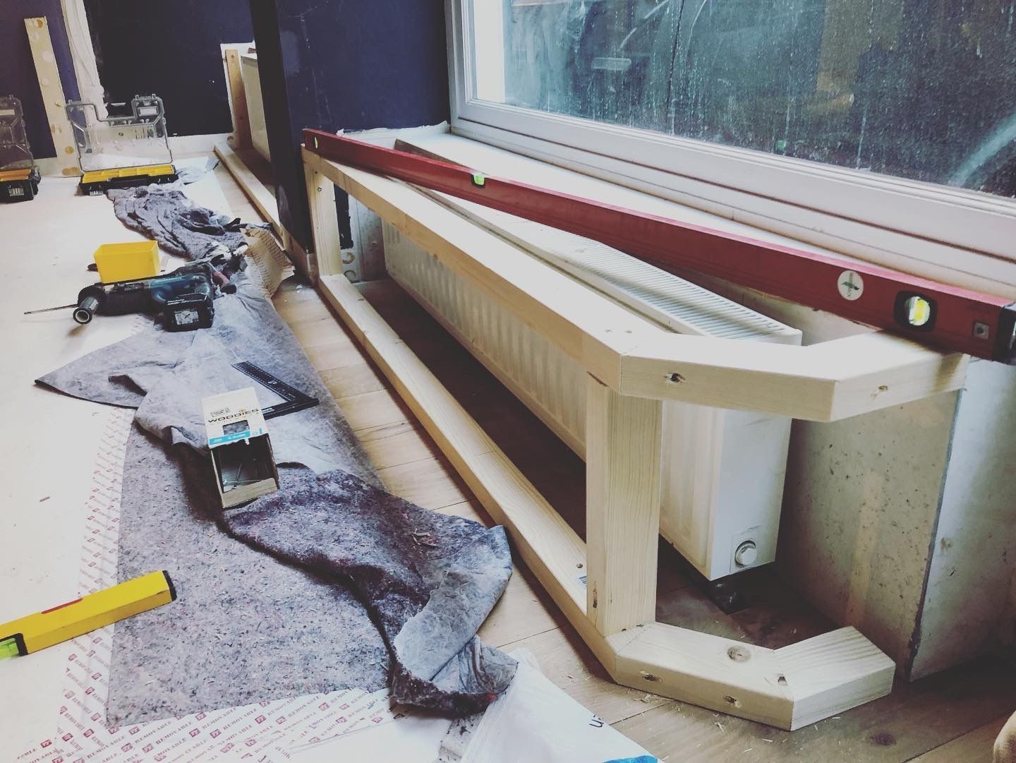 Houten frame rond een radiator onder een raam; gereedschap, waterpas en grijze mat op een lichtgekleurde ondergrond.