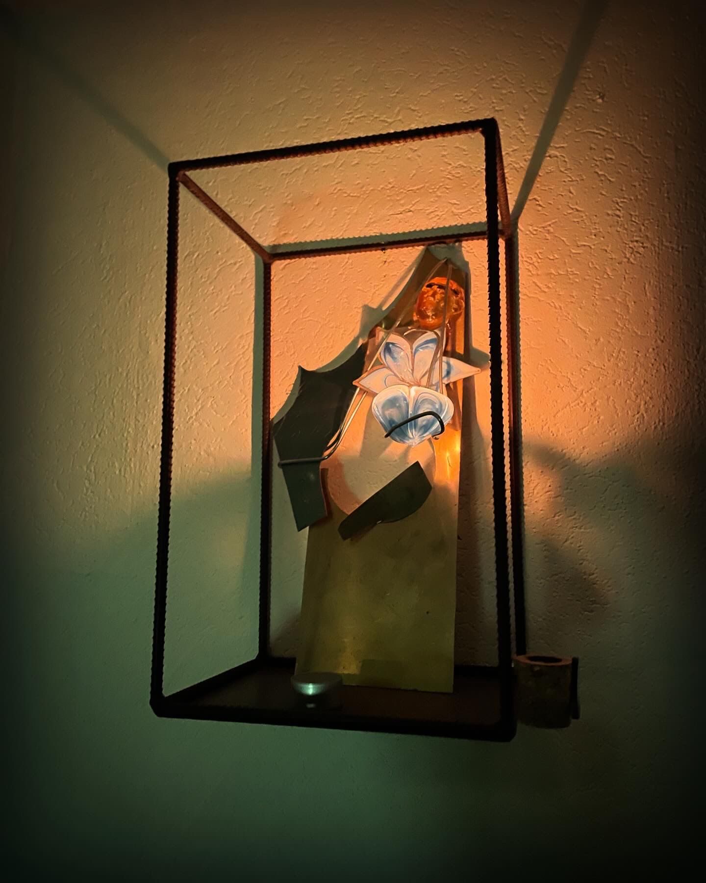 Kunstwerk in een metalen lijst: een mensachtige figuur met blauwe en witte details, tegen een gele achtergrond.