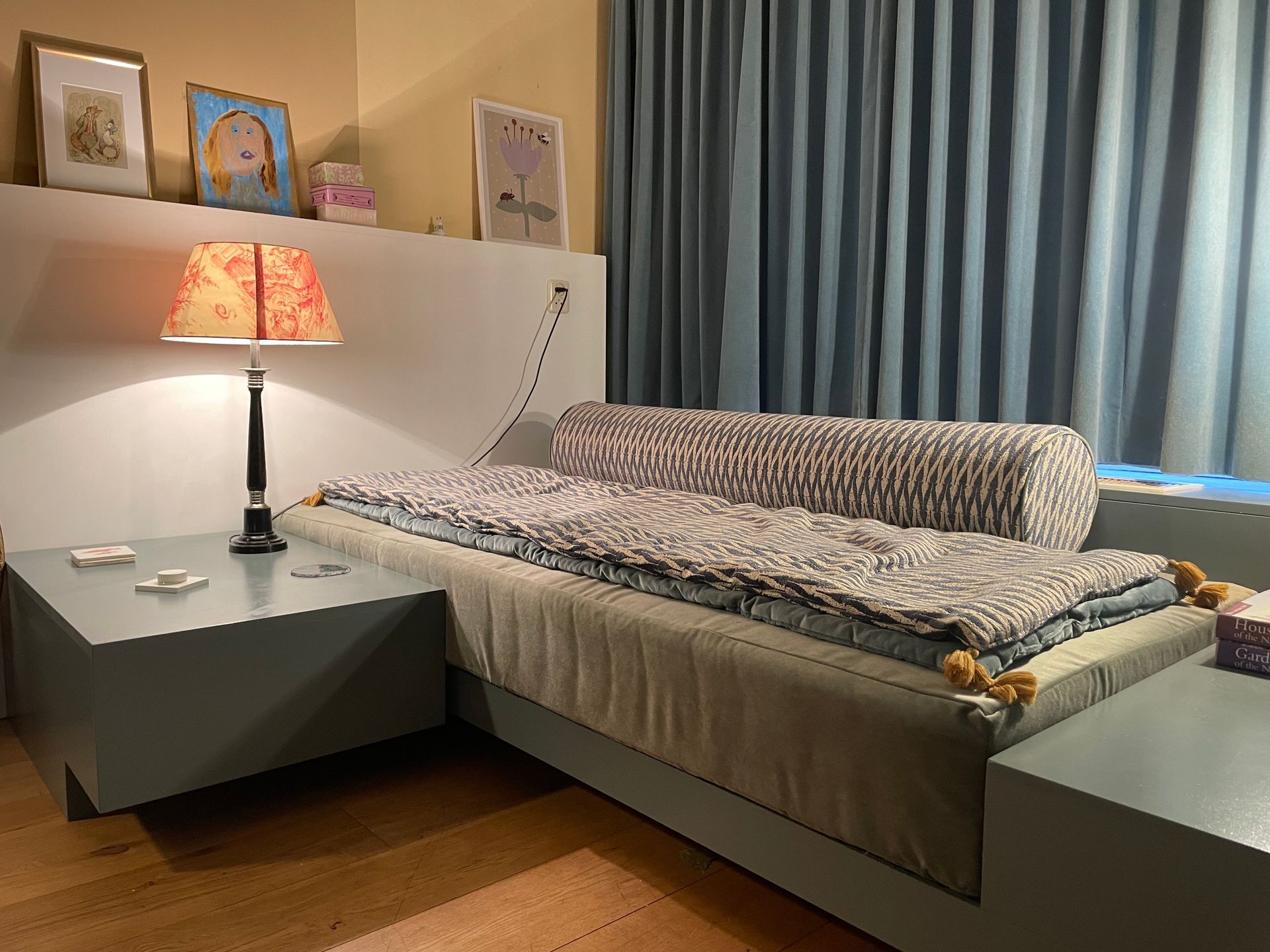 Bed met ingebouwde nachtkastjes en lamp, in een kamer met een raam en blauwe gordijnen.