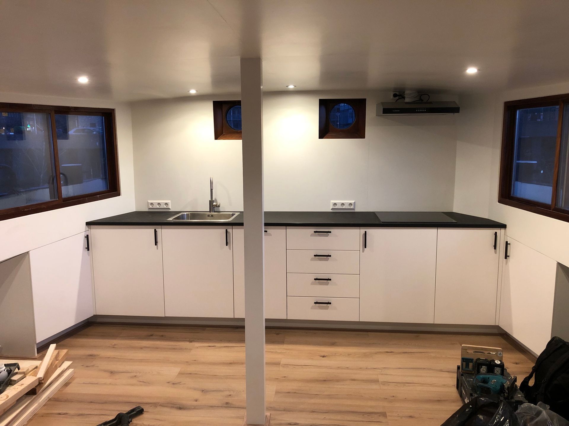 Moderne witte keuken met zwarte aanrechtbladen, houten vloeren en ramen.