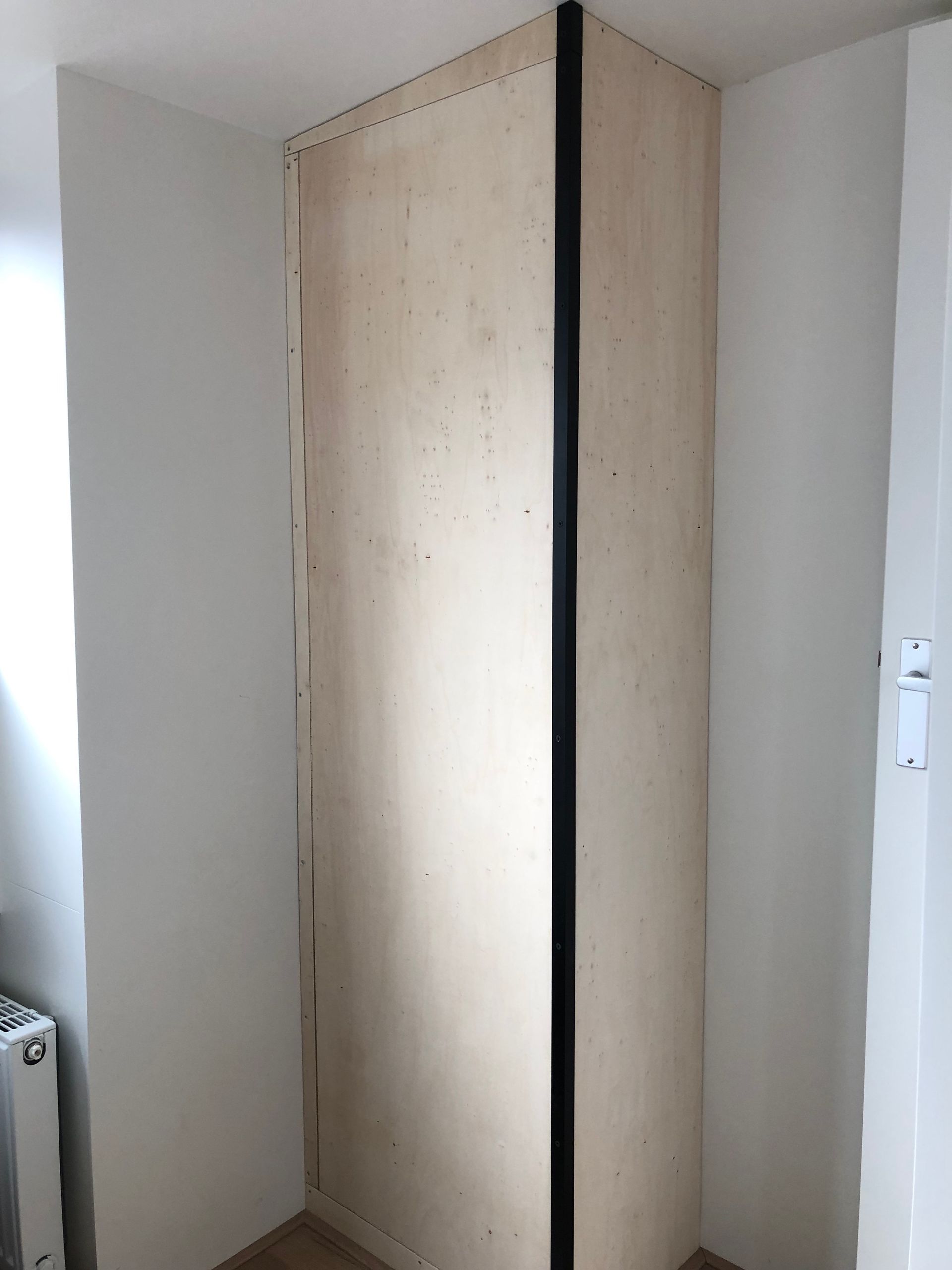 Houten constructie in een hoek. Een zwarte strook scheidt twee houten panelen. Witte muren en deur.