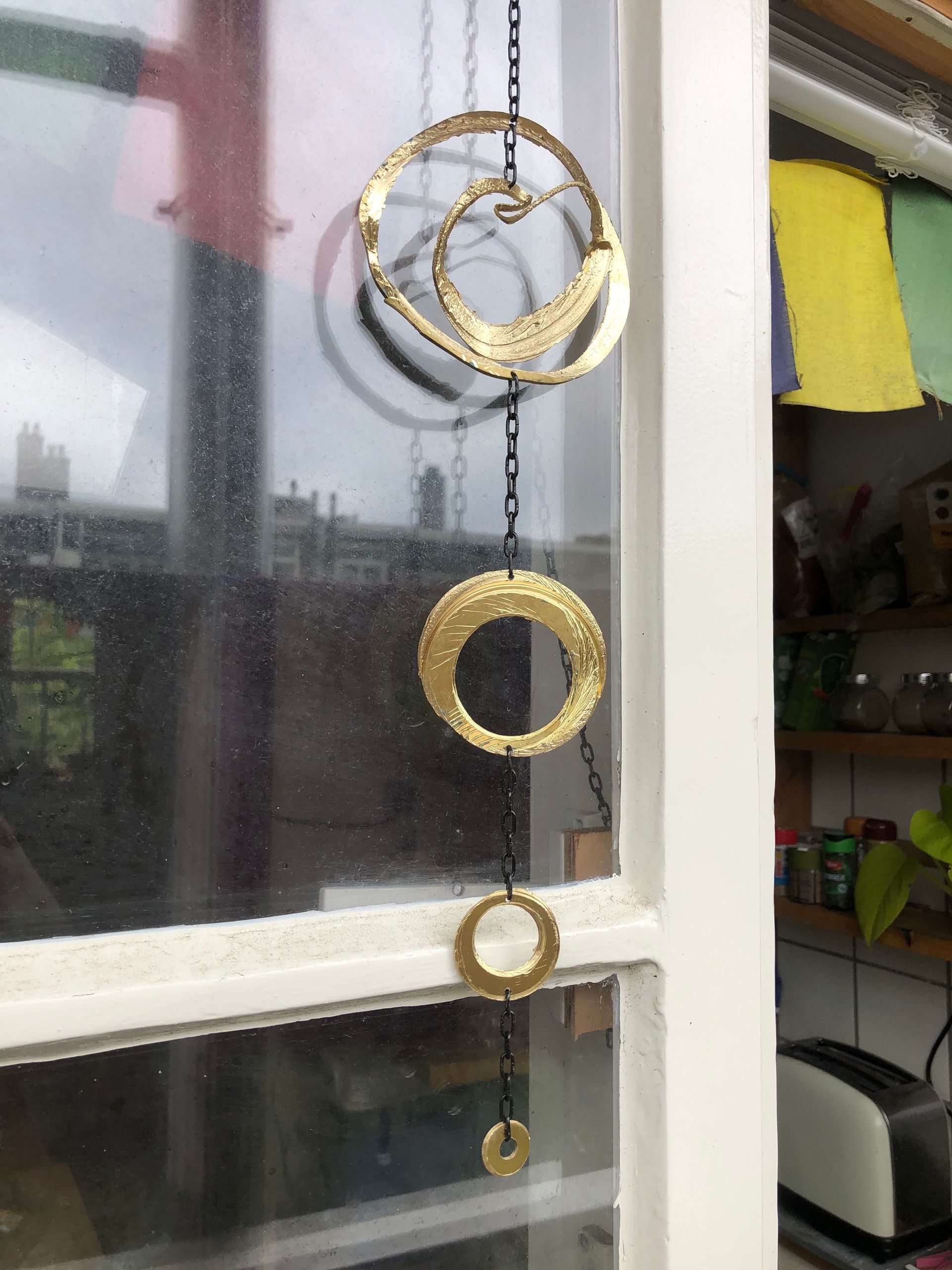 Gouden metalen windgong met cirkel- en halvemaanvormen, hangend in een raam.