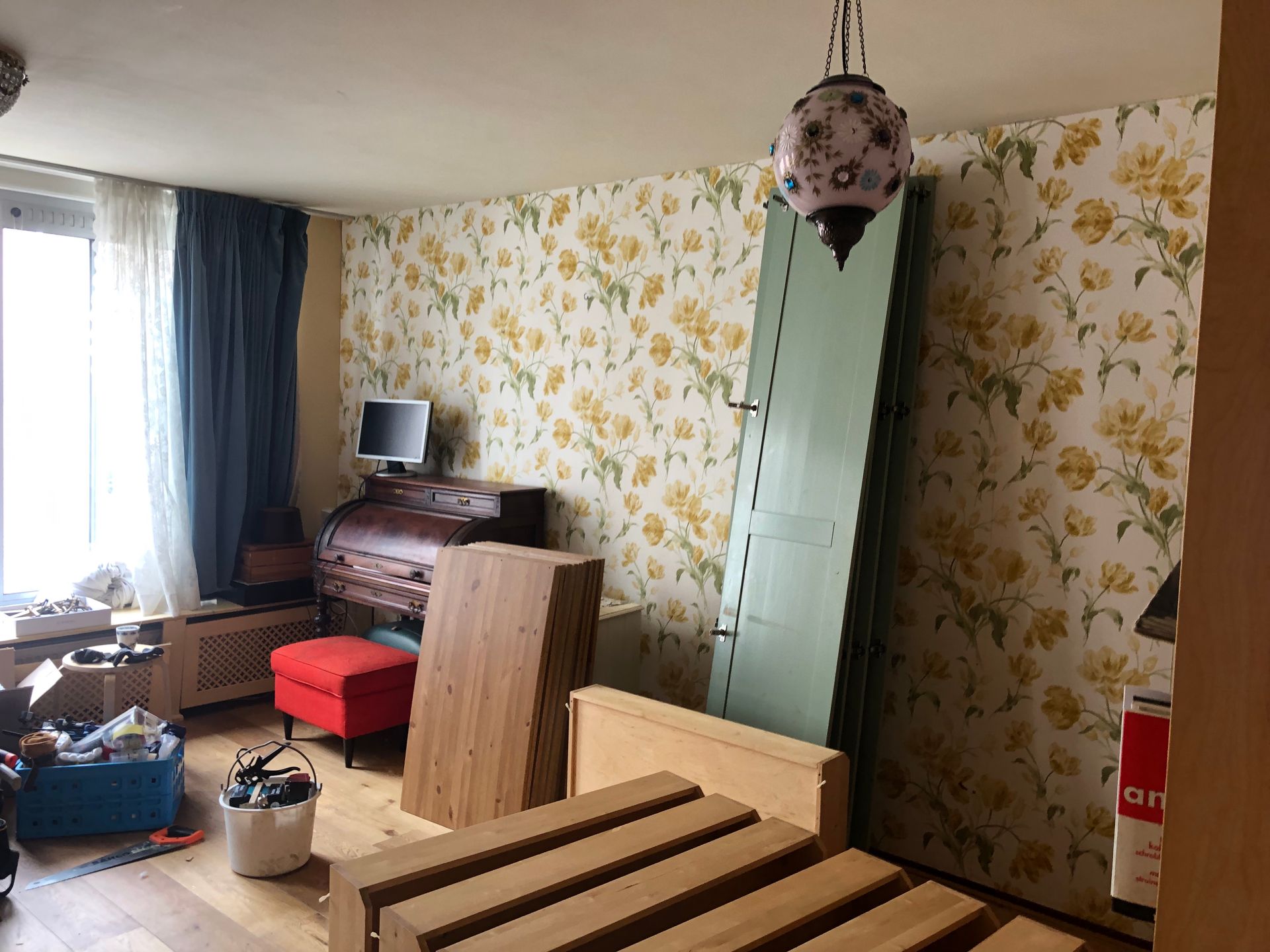 Kamer met bloemenbehang en meubilair, waaronder een hoge groene deur, een bedframe en een commode.