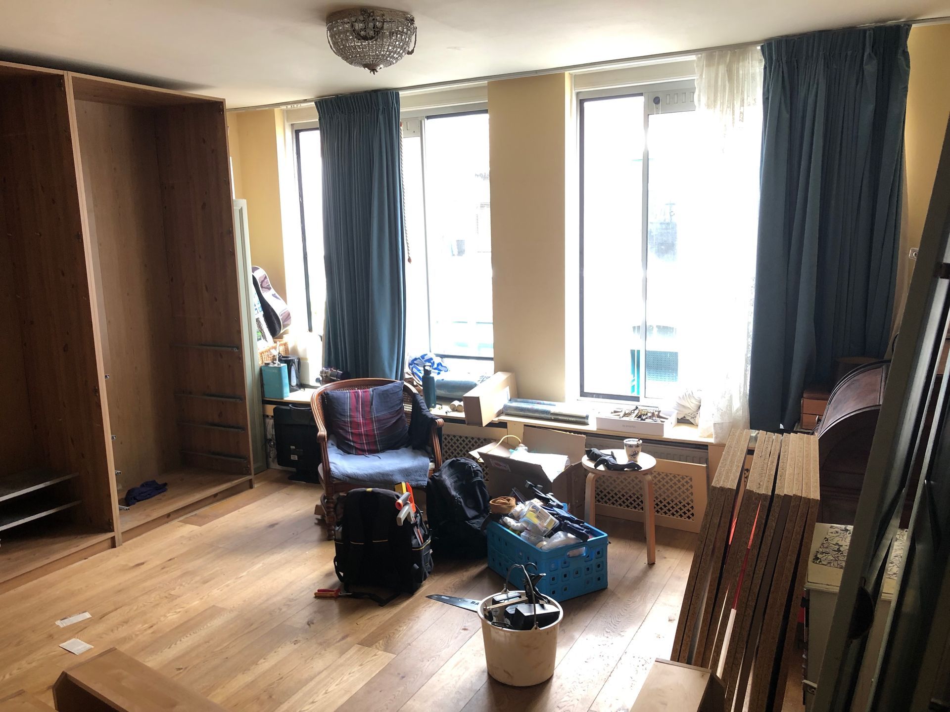 Lege kamer met houten vloer, ingebouwde kast, ramen met blauwe gordijnen en diverse spullen die er rondslingeren.