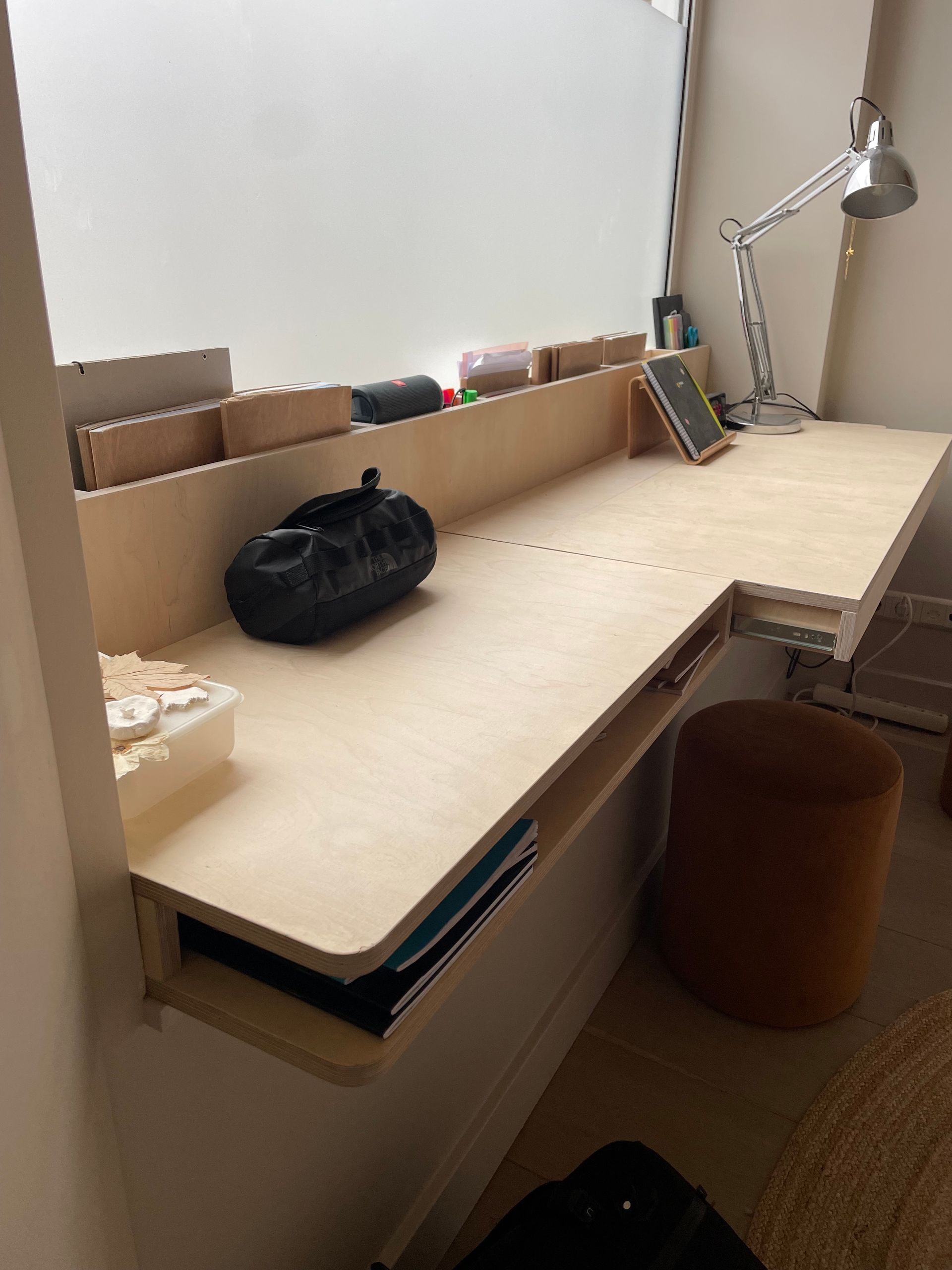 Ingebouwd houten bureau met opbergruimte, tegen een raam. Zwarte tas en lamp erop; krukje ernaast.