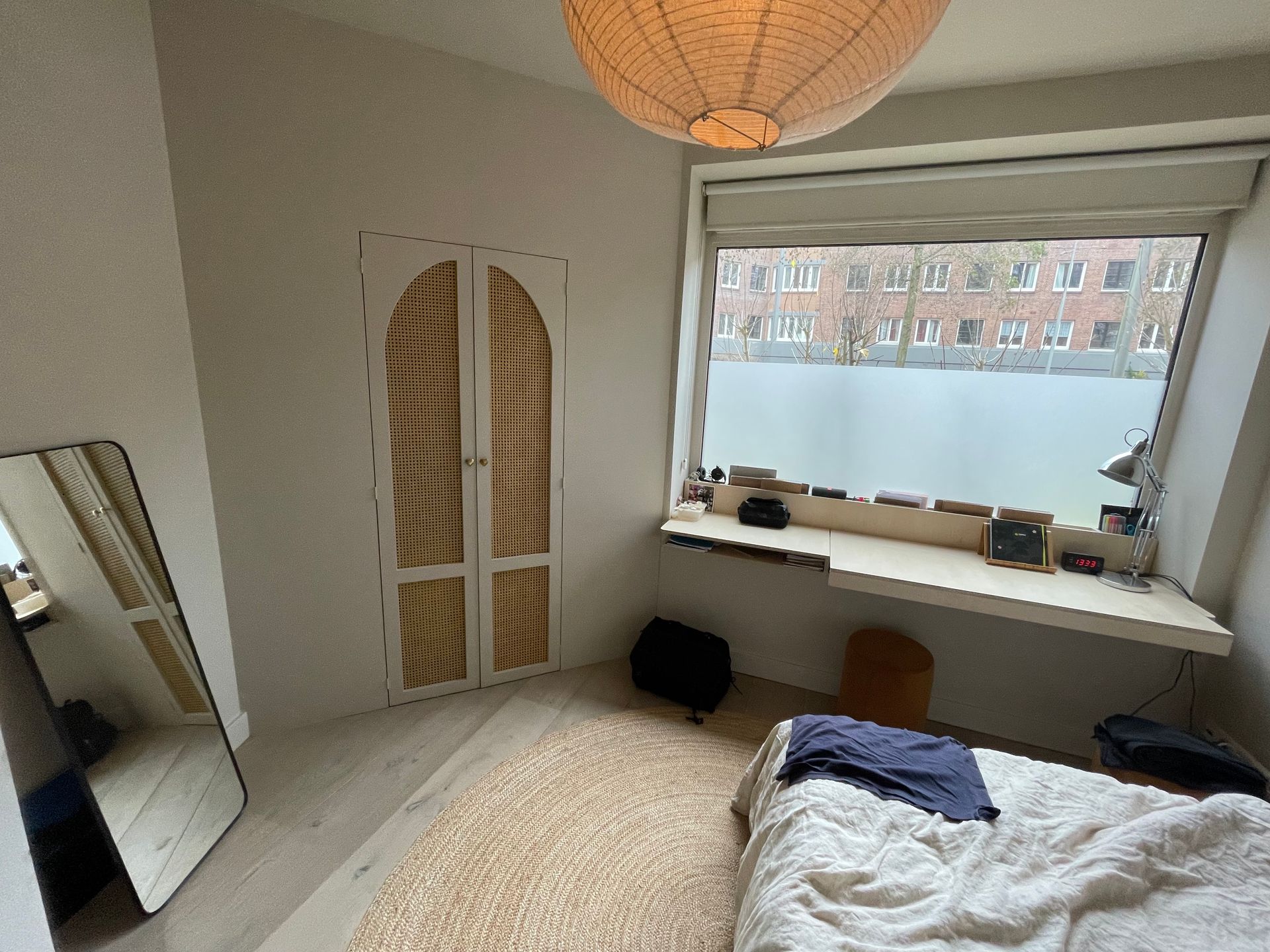 Slaapkamer met groot raam, bed, decoratieve deuren, spiegel, beige vloerkleed en ronde lamp.