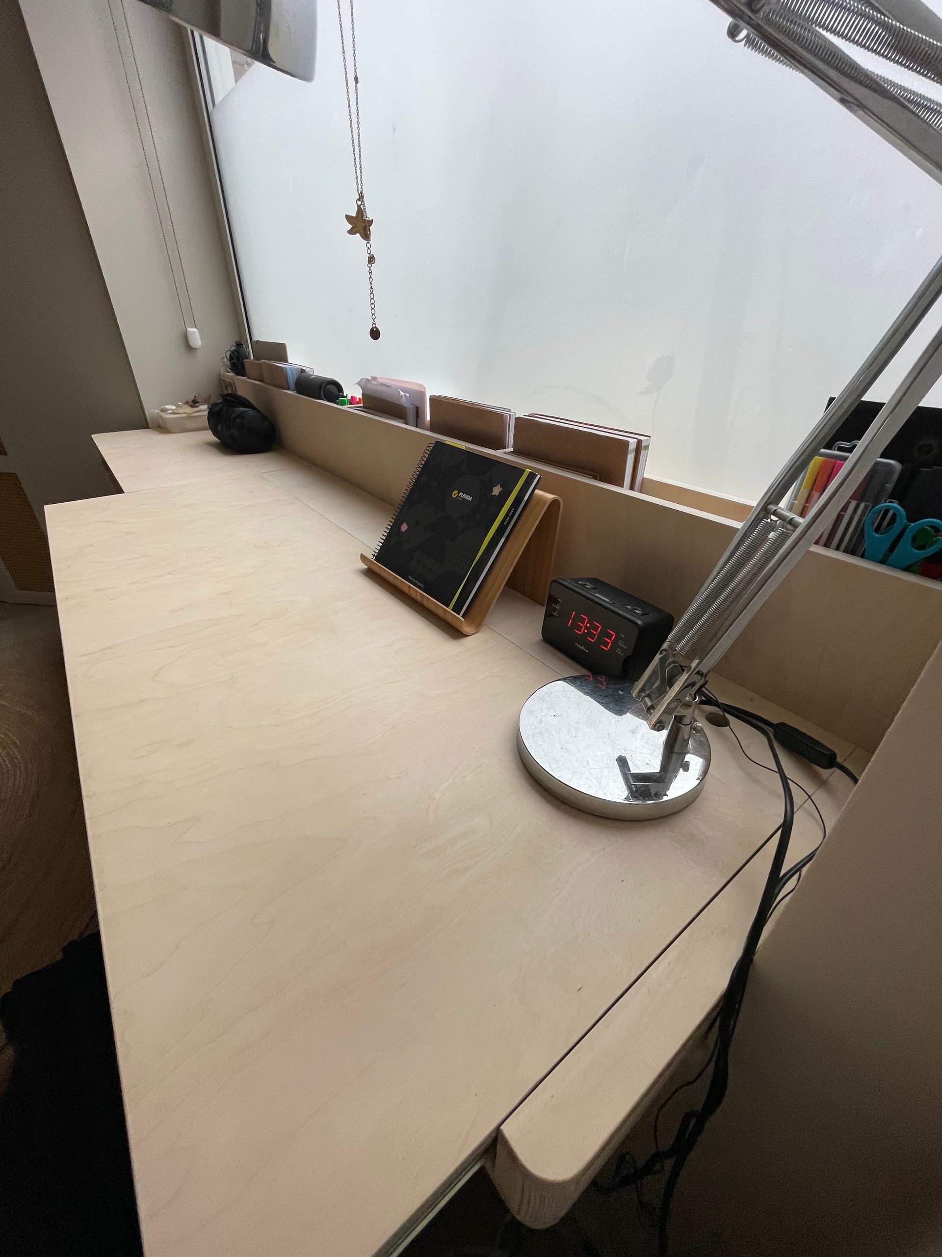 Een licht houten bureau met lamp, boeken en andere voorwerpen bij een raam.
