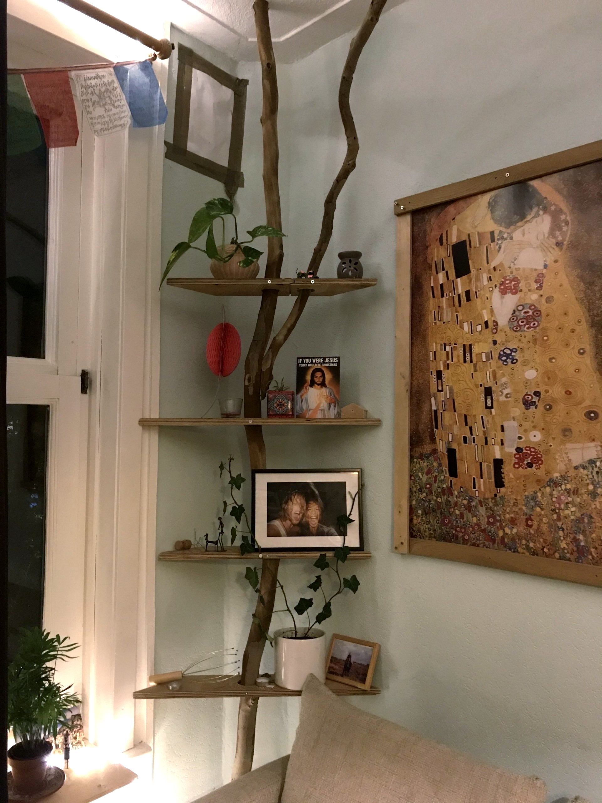 Hoekplanken gemaakt van takken, waarop planten, foto's en decoraties staan; naast een schilderij.