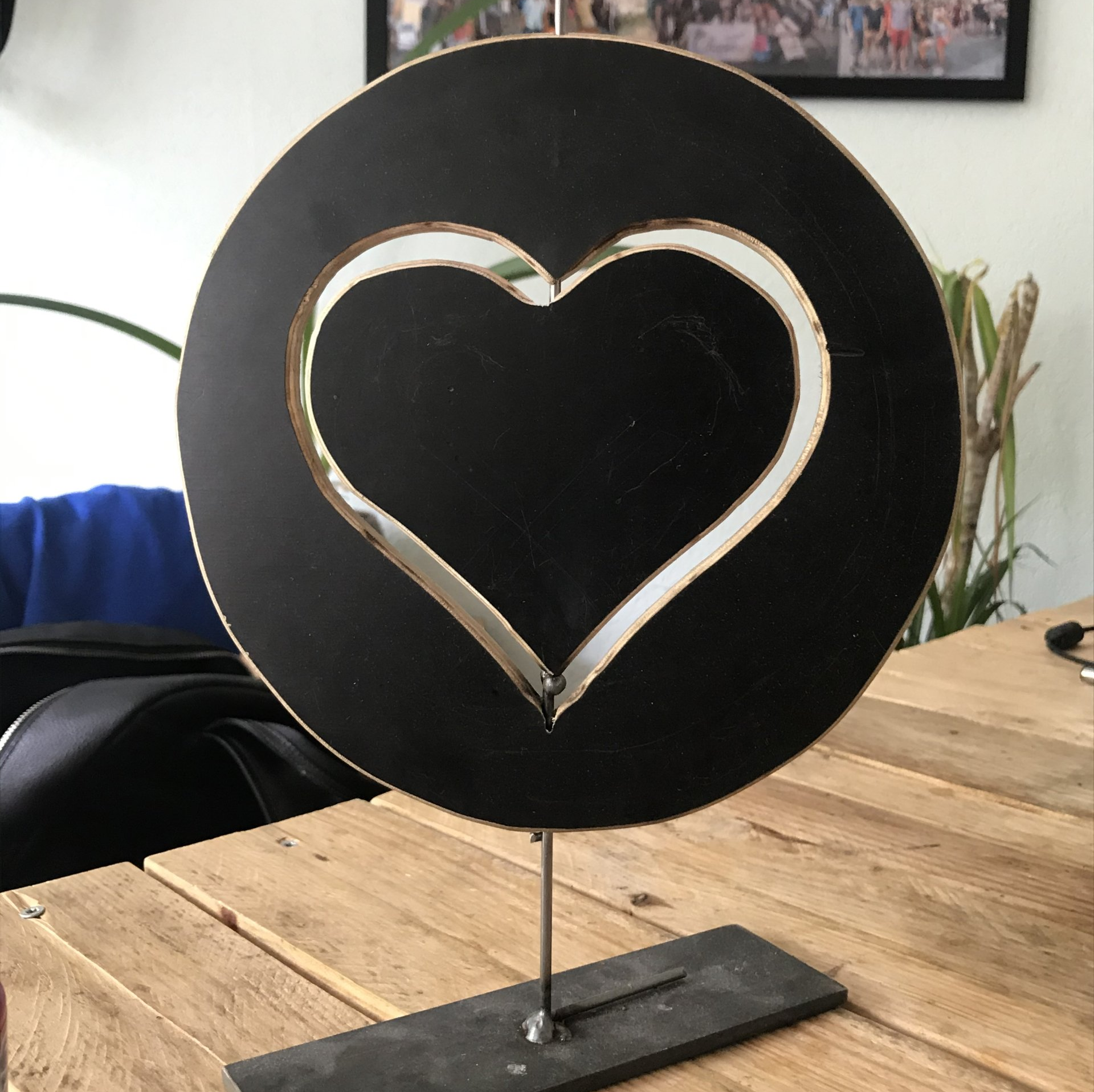 Een zwart hartvormig uitgesneden figuur in een zwarte cirkel, gemonteerd op een metalen standaard, op een houten tafel.