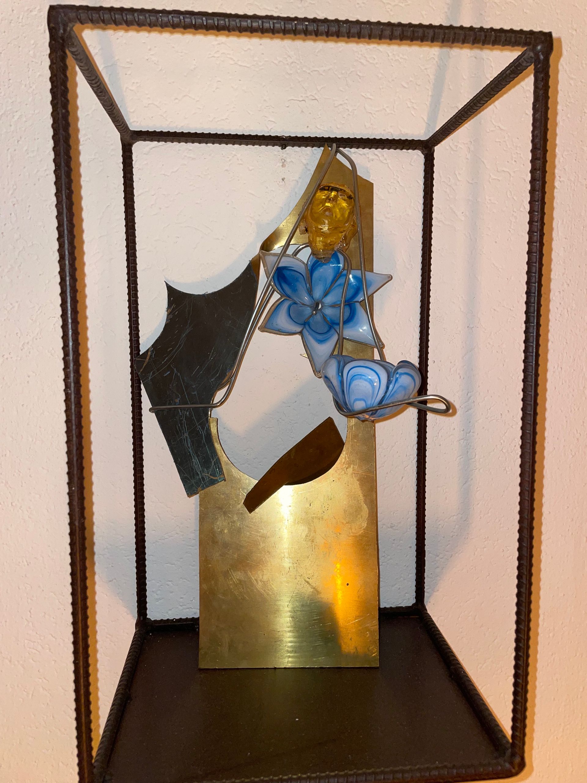 Messing en stalen sculptuur met een blauwe bloem in een donkere metalen vitrine.